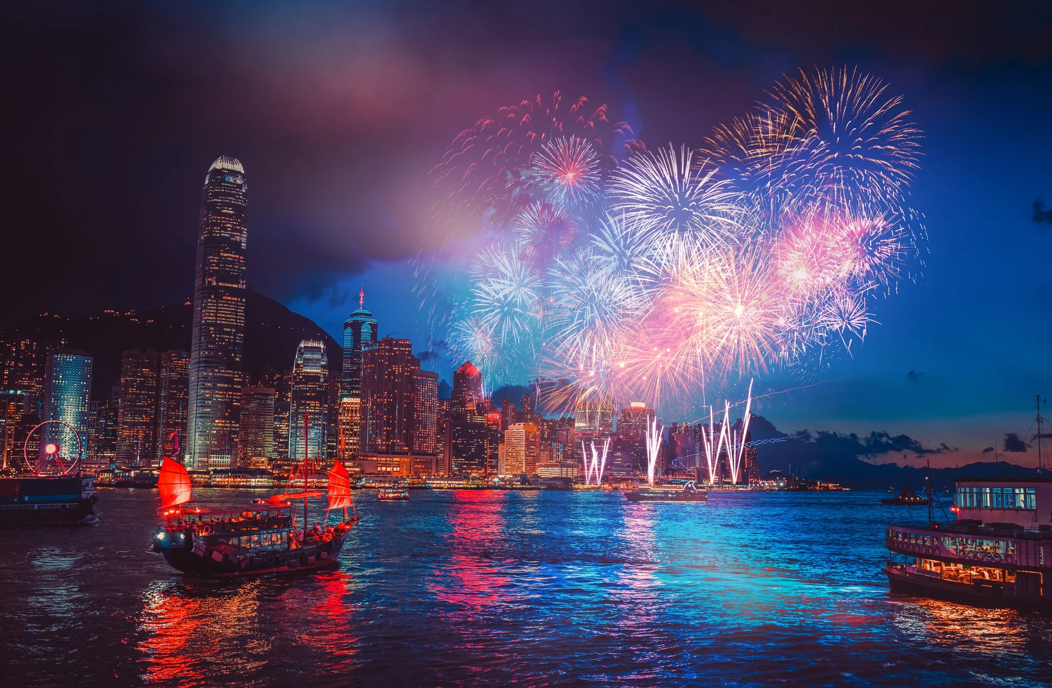 Nighttime city skyline with fireworks display over the water, with boats and sailboats lit by red lights in the foreground.