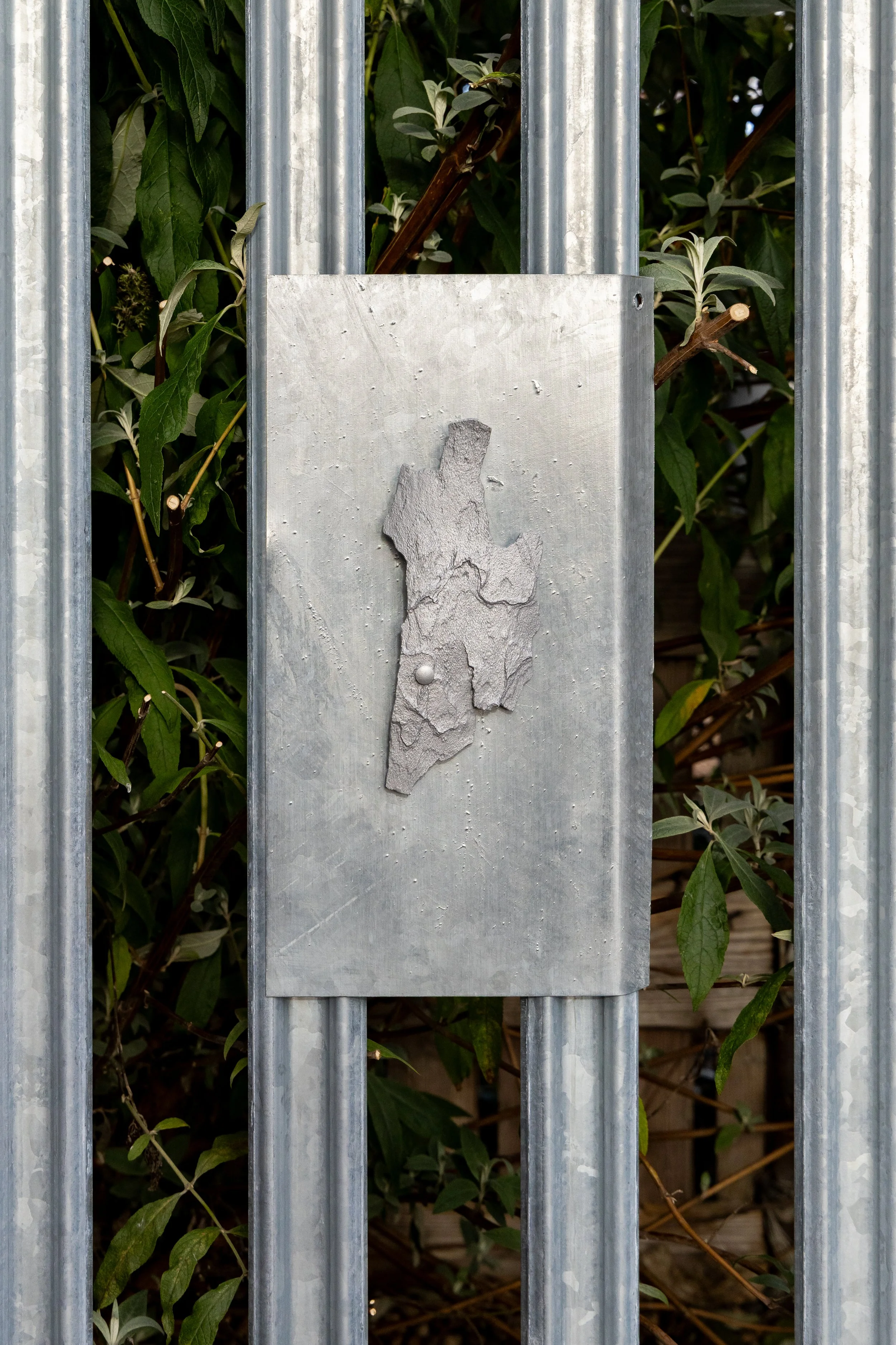 Metal fencing with zinc cast of tree bark attached, with green leaves and branches visible behind and between the bars.