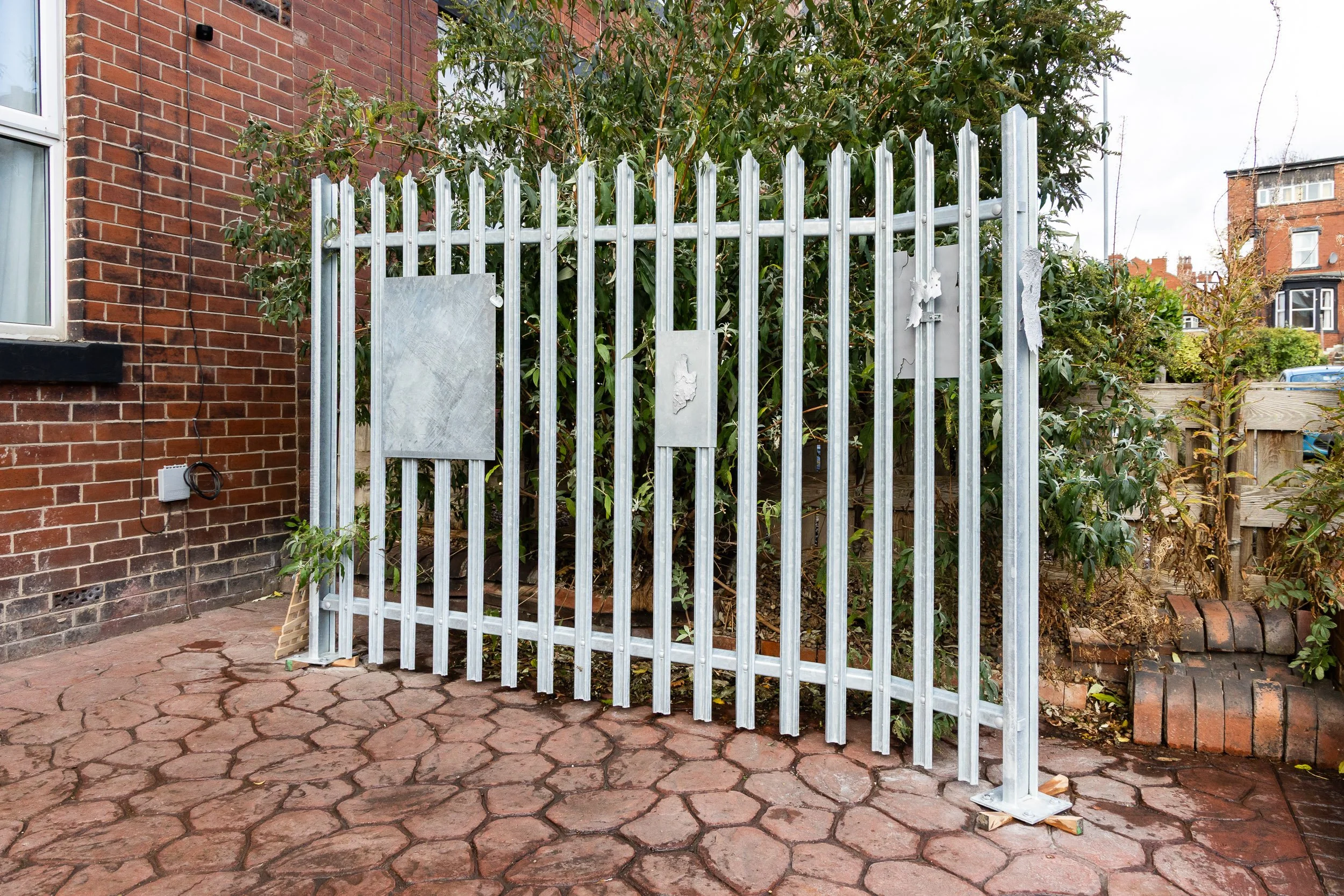 A metal gate in front of a garden with green bushes and trees, next to a brick house wall with a window and electric wiring.