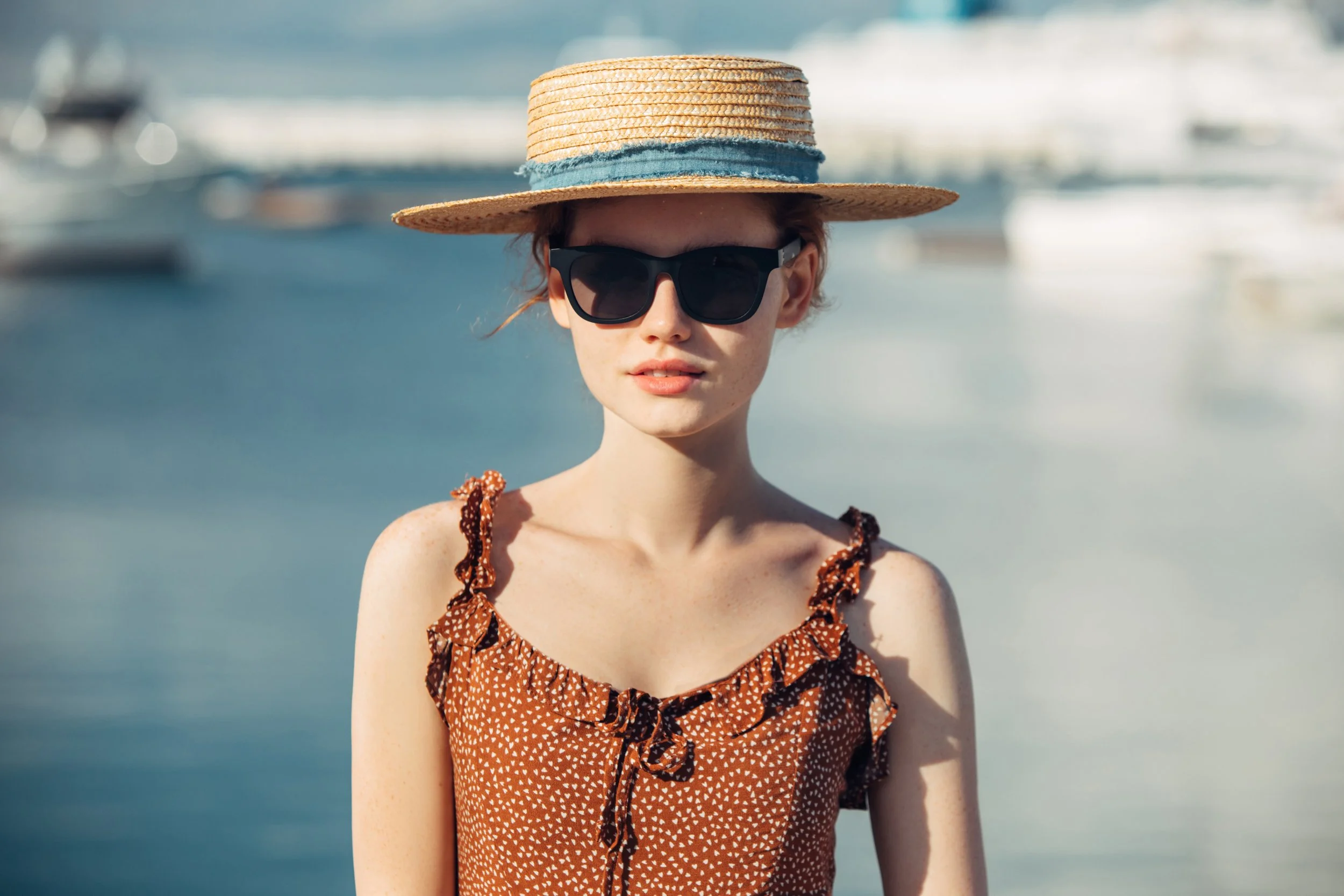 Woman wearing a brown summer sundress, straw hat and sunglasses by the water in Mediterranean vacation style.