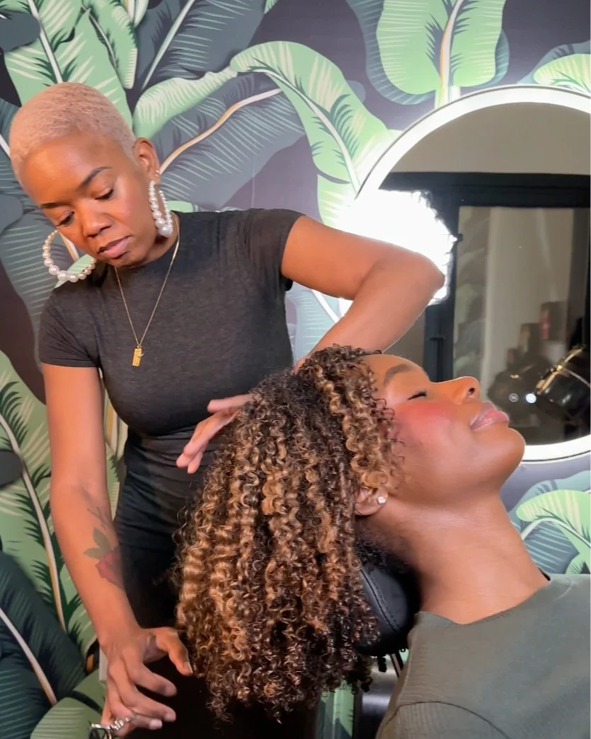 Woman with curly hair receiving a scalp massage at beauty salon, tropical leaf wallpaper in background.
