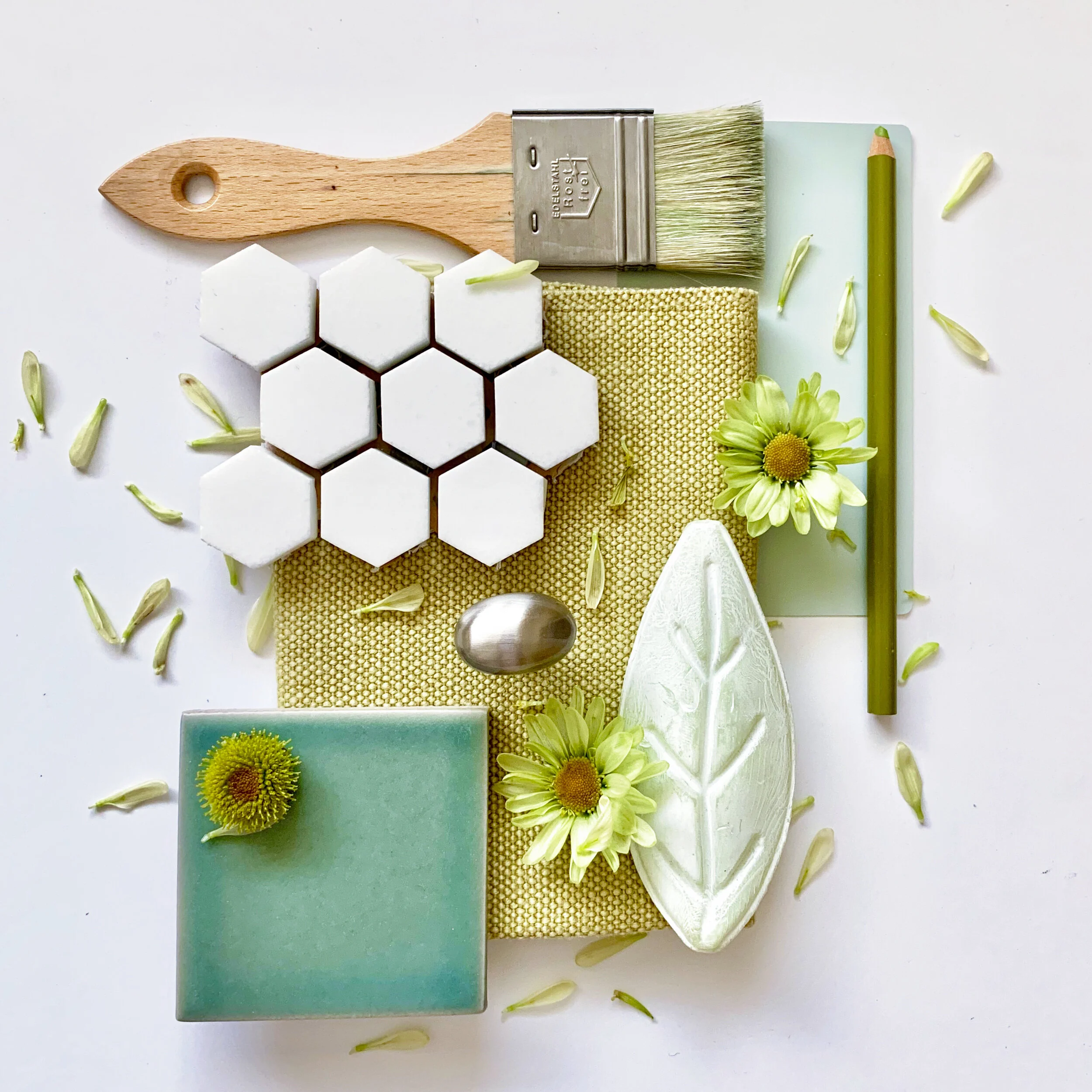 Flat lay of green and white decorative objects, including hexagon tiles, a paintbrush, a green pencil, flowers, leaves, a spoon, and candles on a white background.