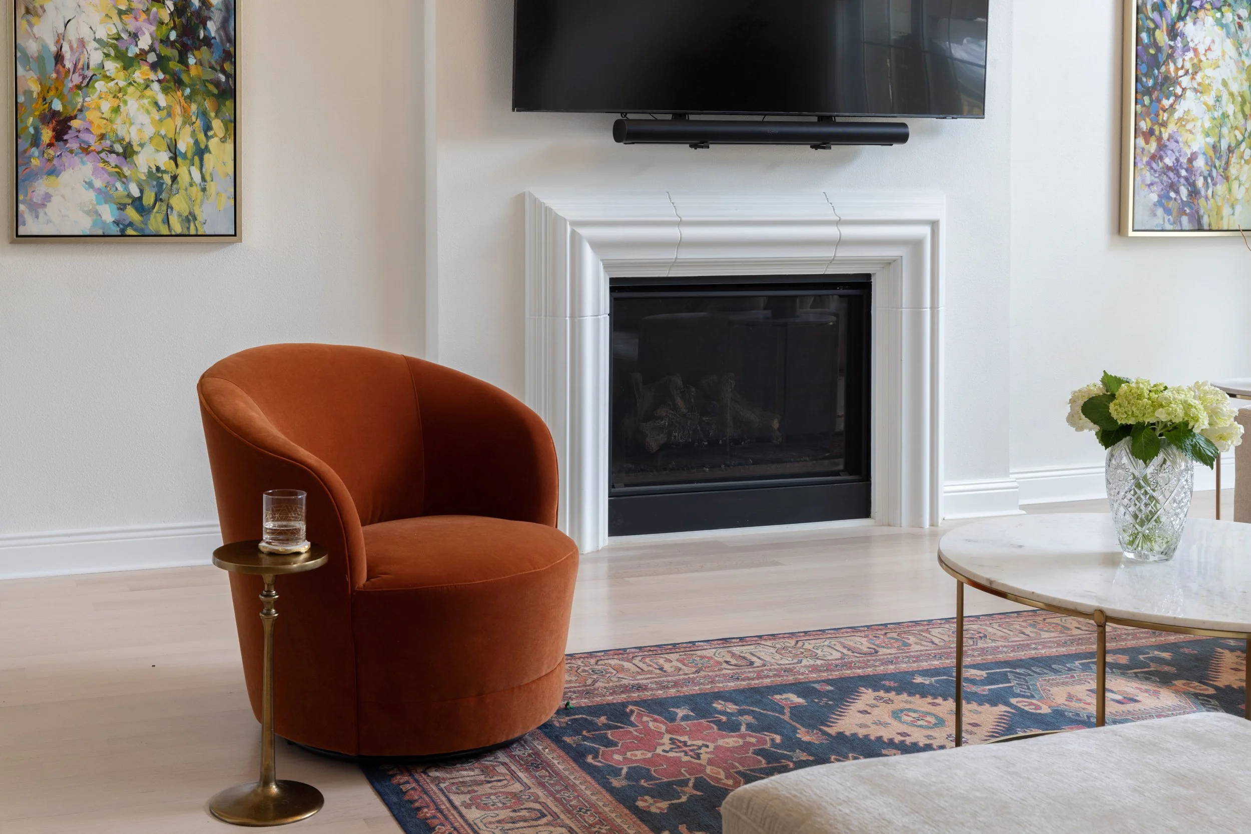 Living room with orange velvet armchair, marble coffee table with a vase of white and green flowers, a fireplace, a wall-mounted TV, and colorful abstract paintings.