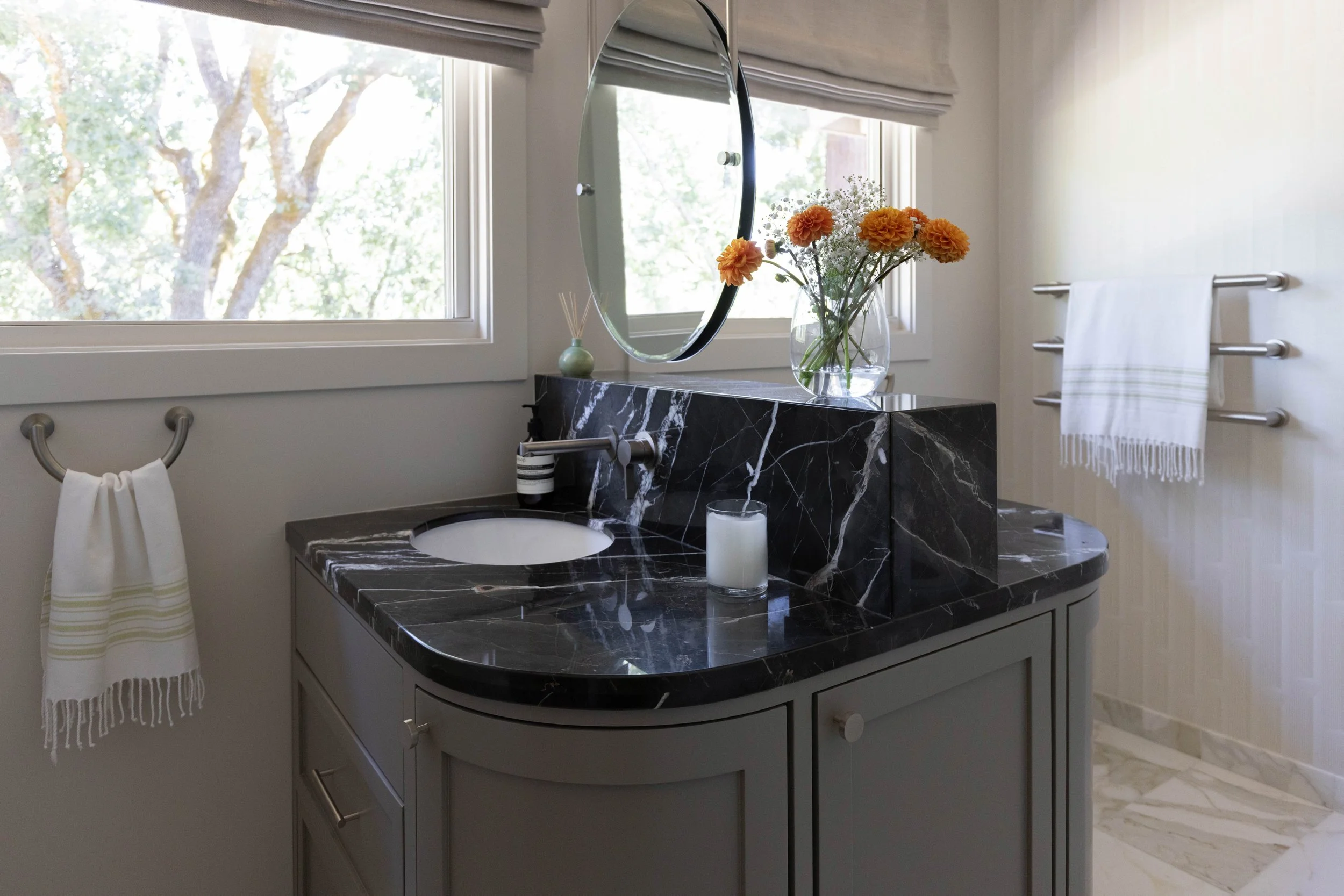 Bathroom with a black marble countertop vanity, a round mirror, a vase of orange flowers, two white towels on towel racks, and a window showing trees outside.