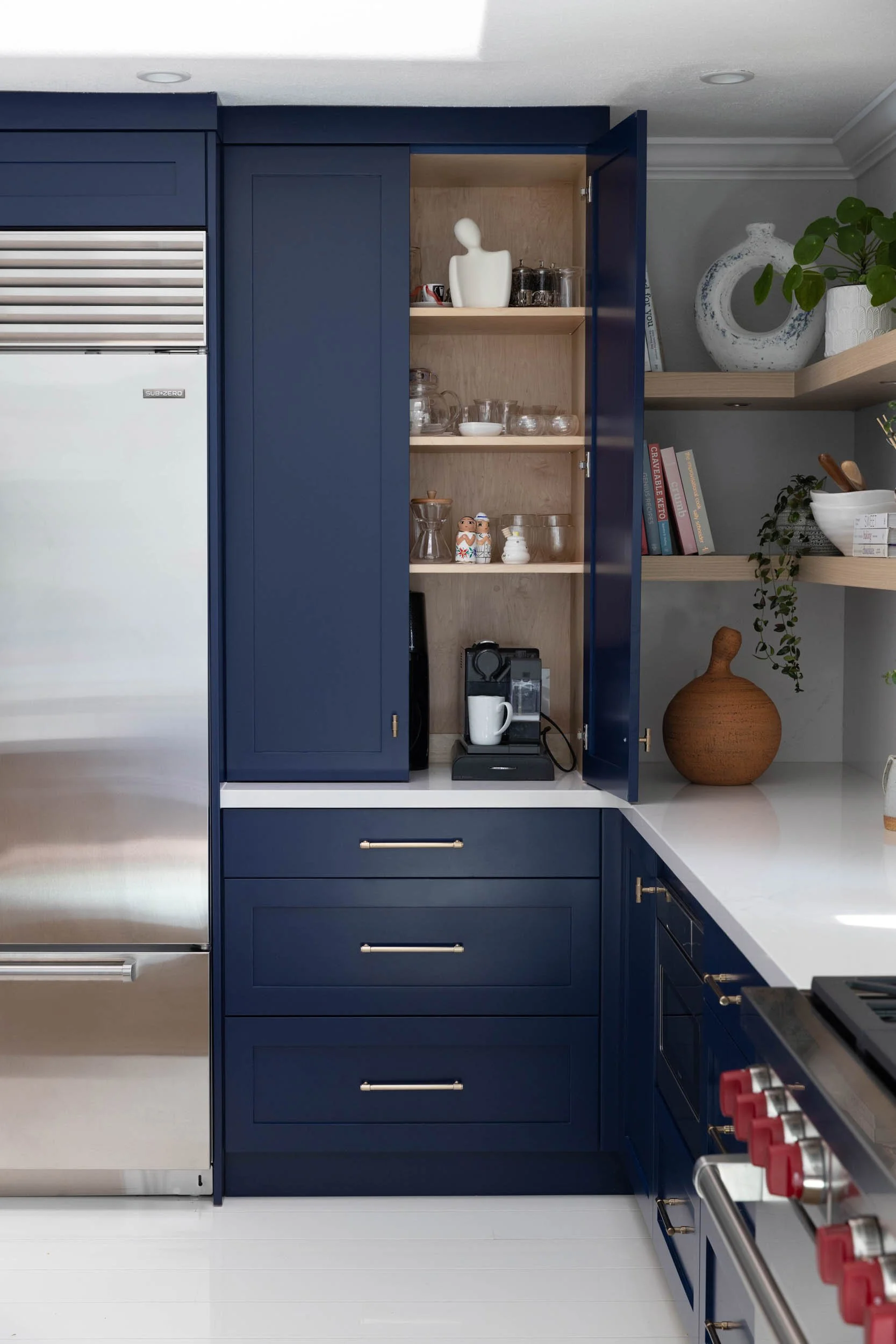 Open navy blue kitchen cabinet filled with glassware and decorative items, adjacent to a white countertop and a Sub-Zero refrigerator.