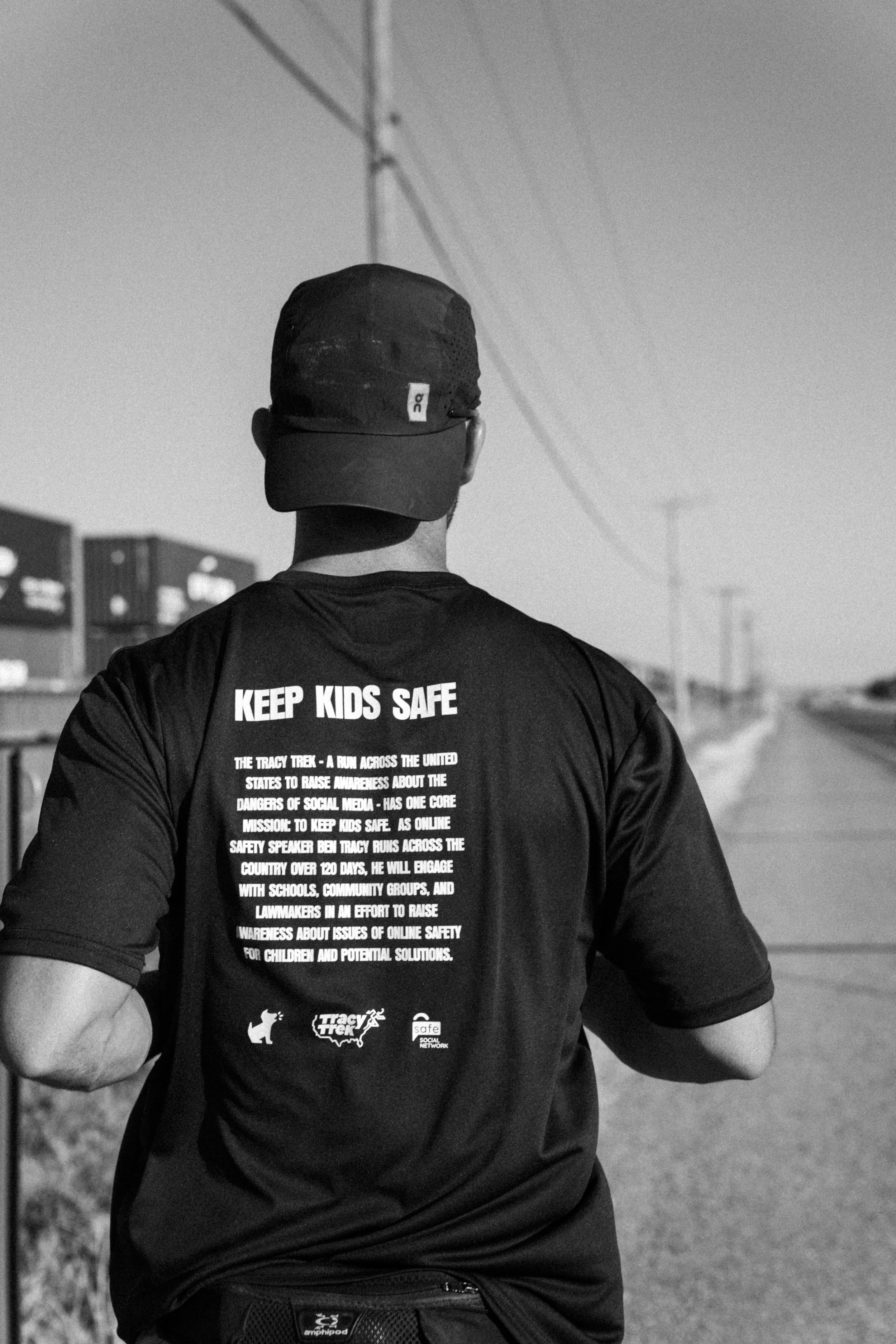 Ben Tracy is running across America with a shirt that has "Keep Kids Safe" printed on the back, running on the side of the road as a train passes.