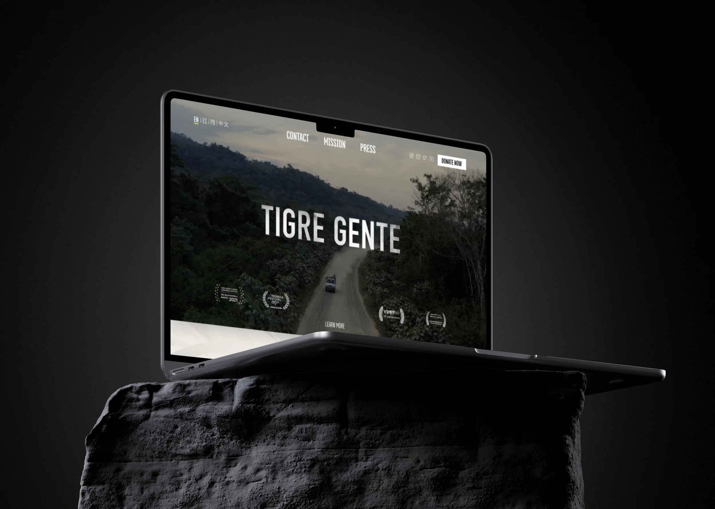 A laptop displaying a website titled 'Tigre Gente' with an image of a forested mountain road, placed on a rough stone surface against a dark background.