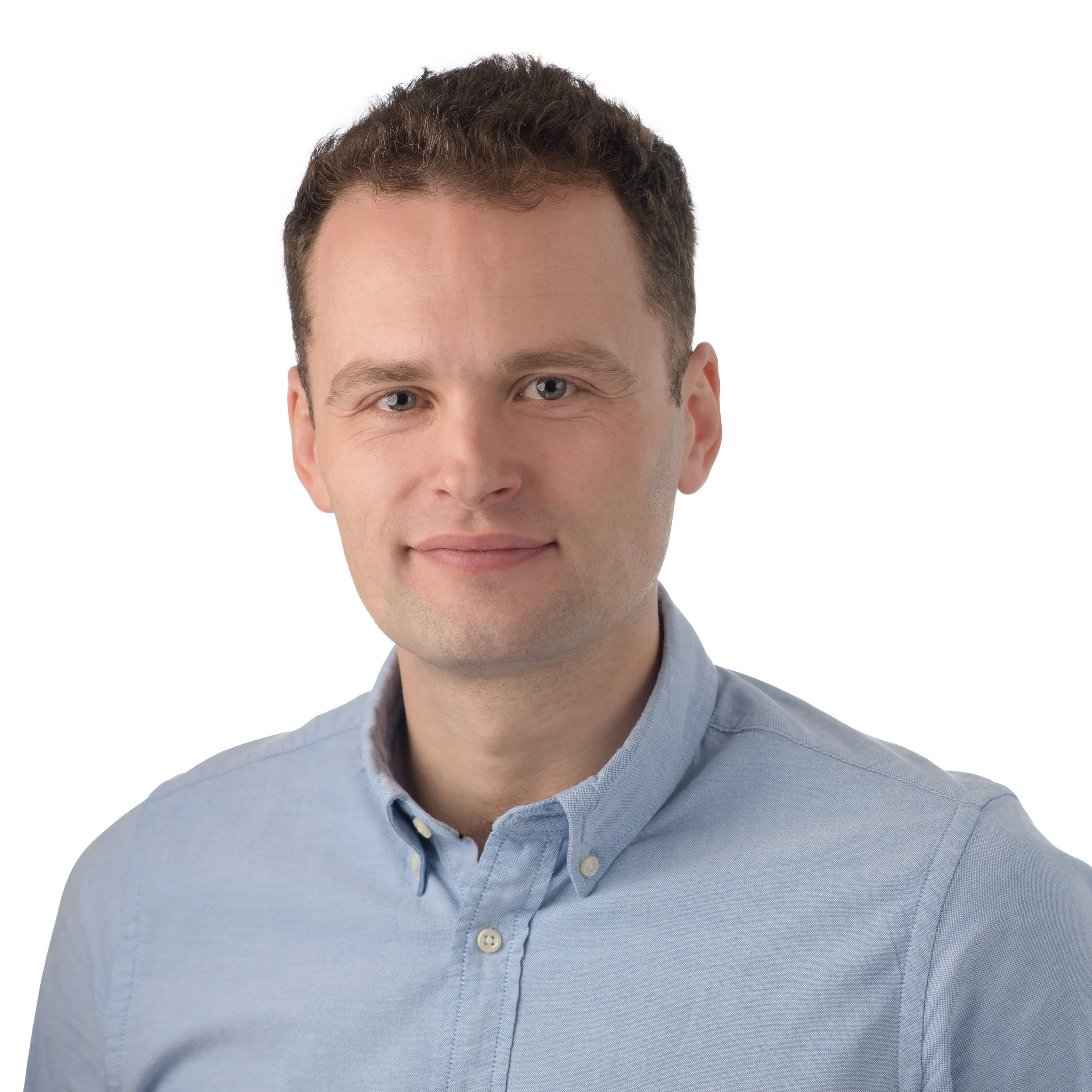 Portrait of a young man with short brown hair wearing a light blue button-up shirt, smiling softly against a white background.