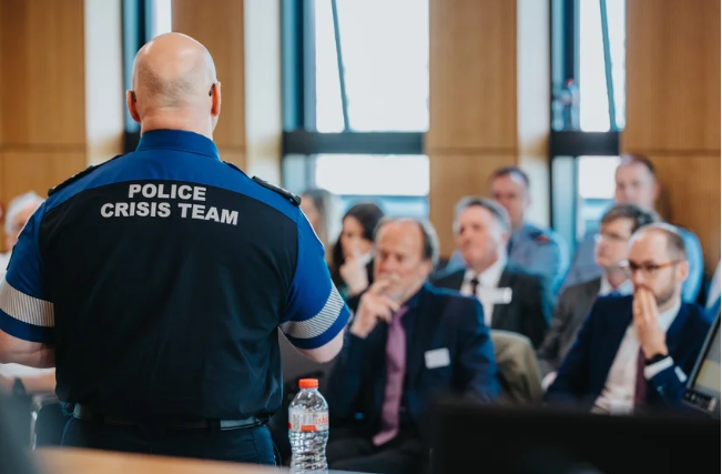 A police crisis team officer speaking to a group of business professionals in a modern conference room.
