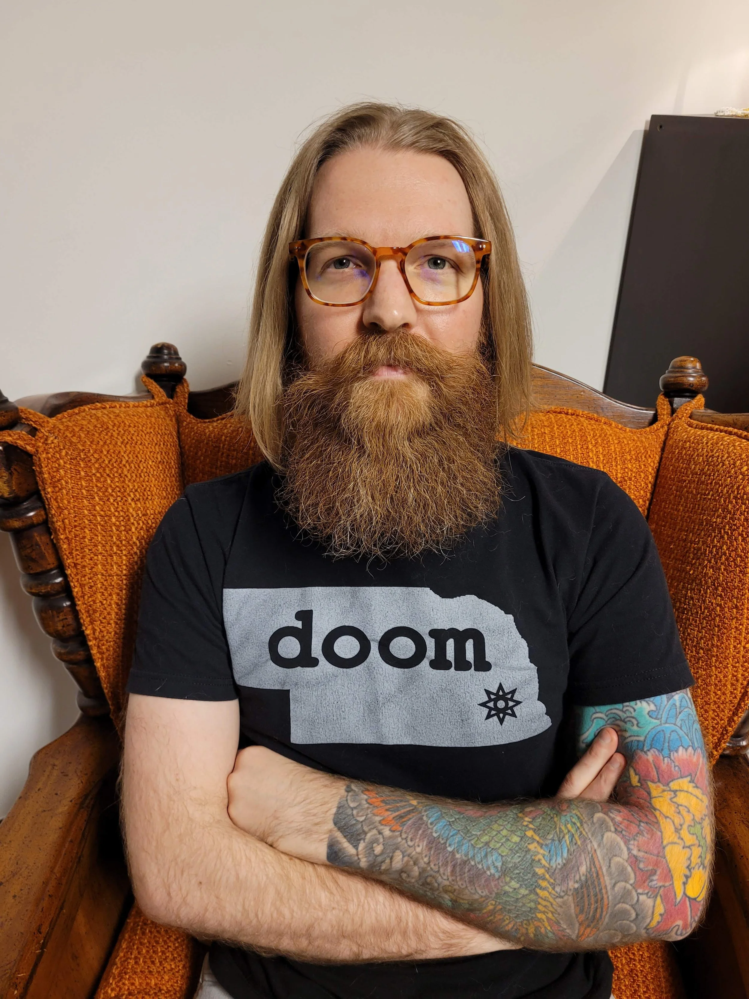 Author Michael Bettendorf sits in a burnt-orange chair. He has shoulder length hair, wears orange tortoise shell glasses and a T-shirt that says "doom" inside the state outline of Nebraska.