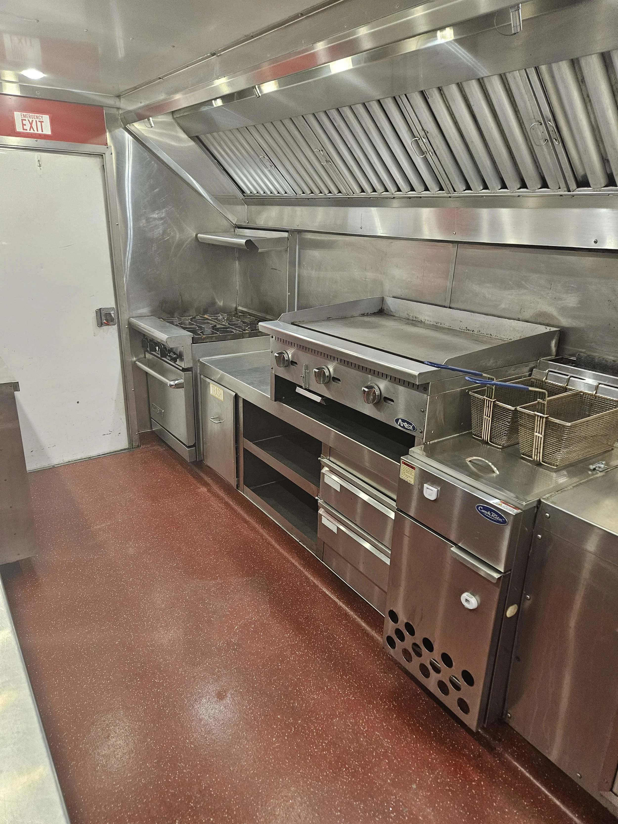 Commercial kitchen with stainless steel appliances, overhead exhaust hood, gas stove, flat grill, fryer baskets, and storage drawers, with a red speckled non-slip floor.