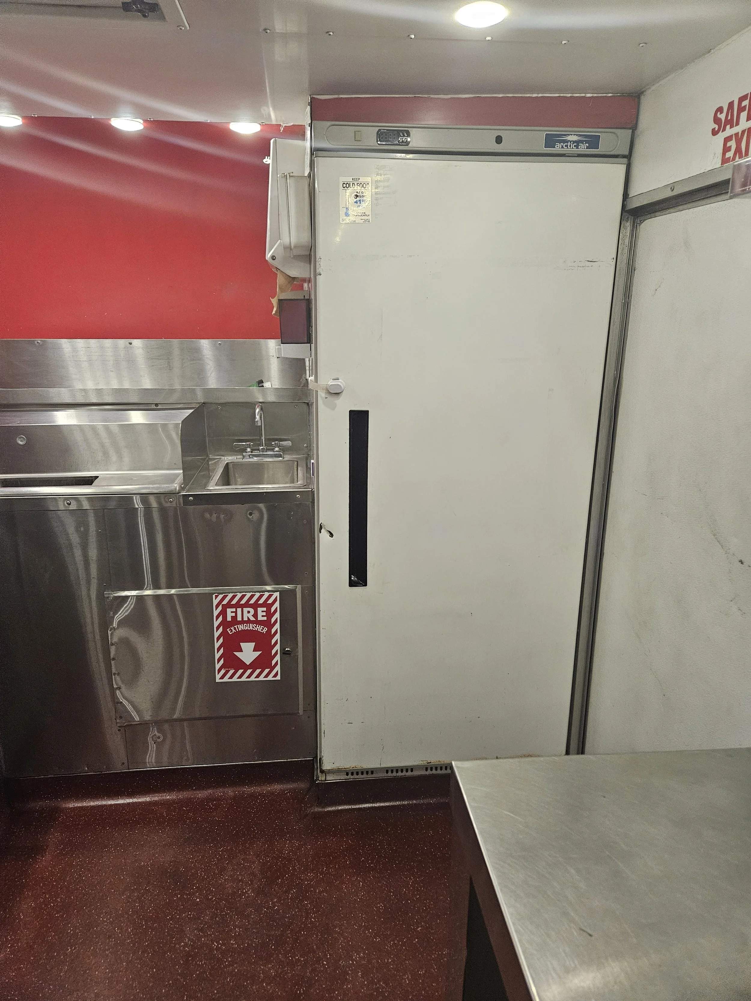 A commercial kitchen with stainless steel sink and counter, white door with black handle, and red fire extinguisher sign on the wall.