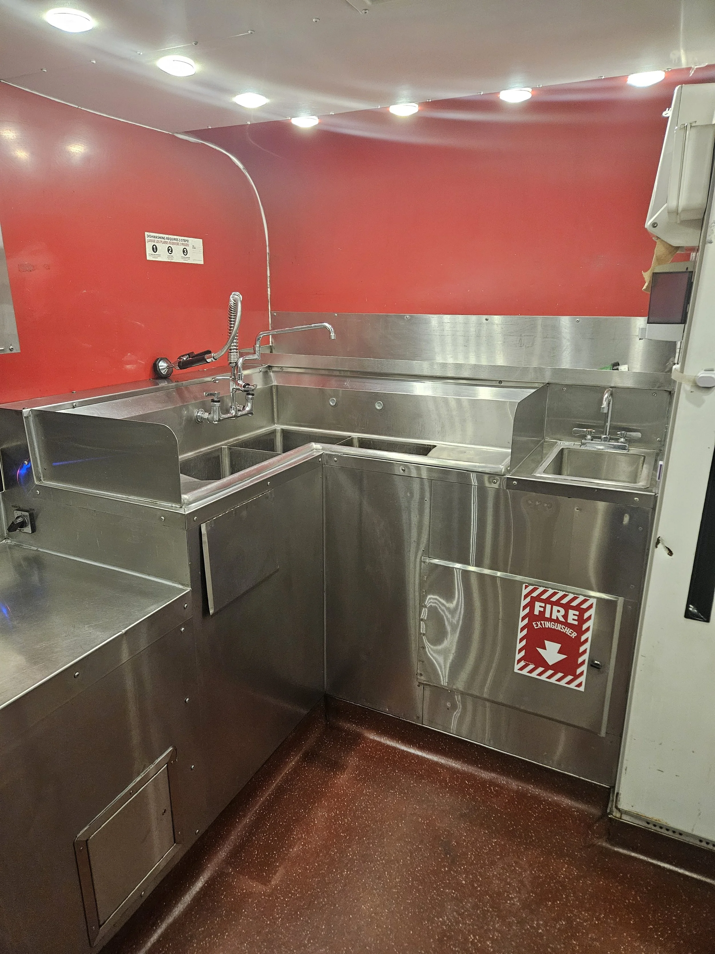 Commercial stainless steel kitchen sink with multiple basins, a faucet with a flexible hose, and a fire extinguisher cabinet in a red wall environment.