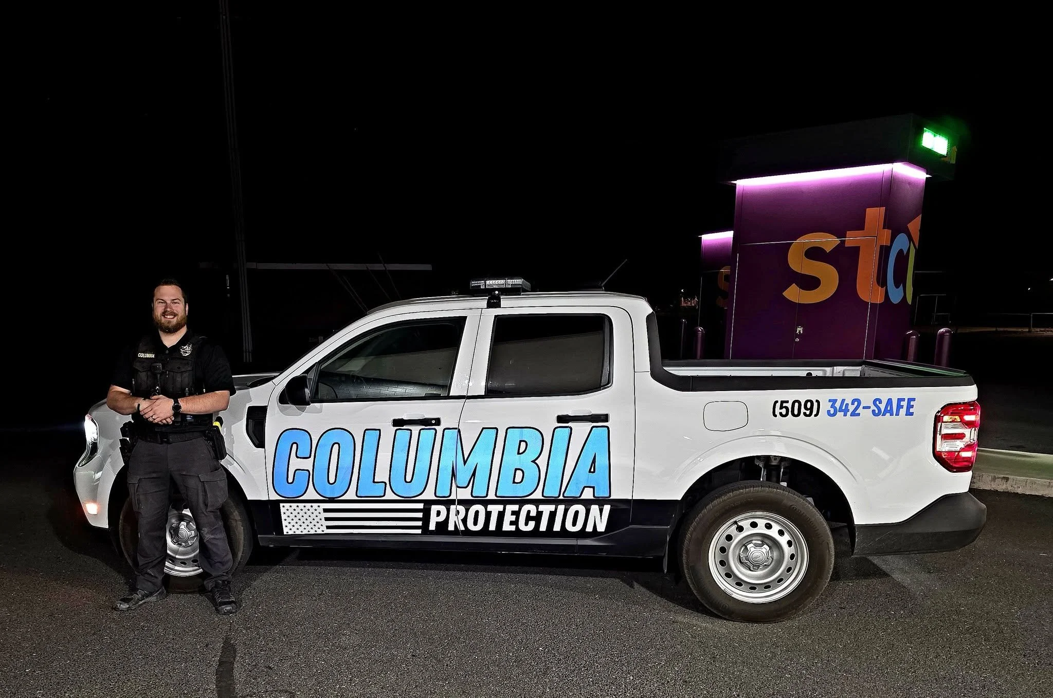 A male officer standing next to a white police vehicle marked 'Columbia Protection' at night. The officer is smiling, wearing black uniform, and the vehicle has a stop sign and a neon lighted building in the background.
