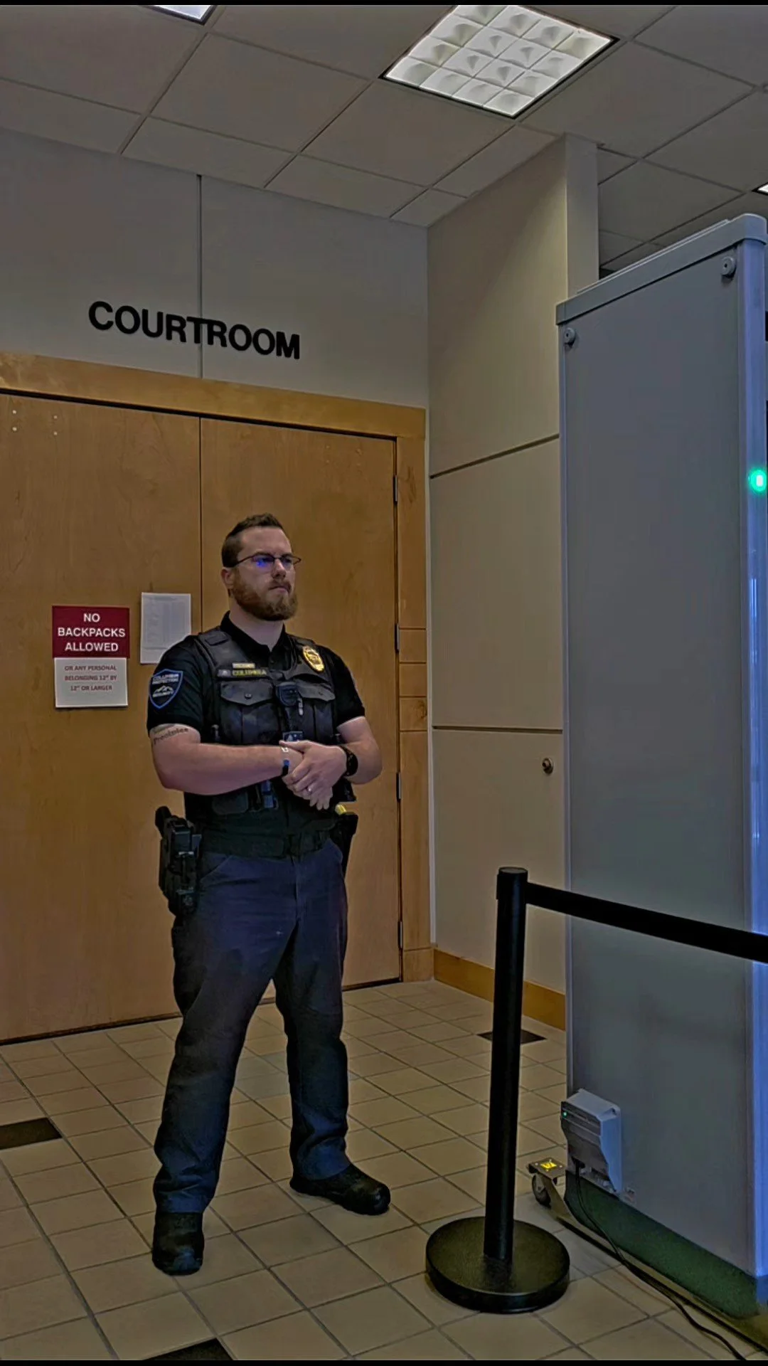 officer standing with arms crossed in front of a courthouse entrance. The officer is wearing a black uniform, glasses, and has a utility belt with a gun holster. A sign on the door reads "No backpacks allowed".