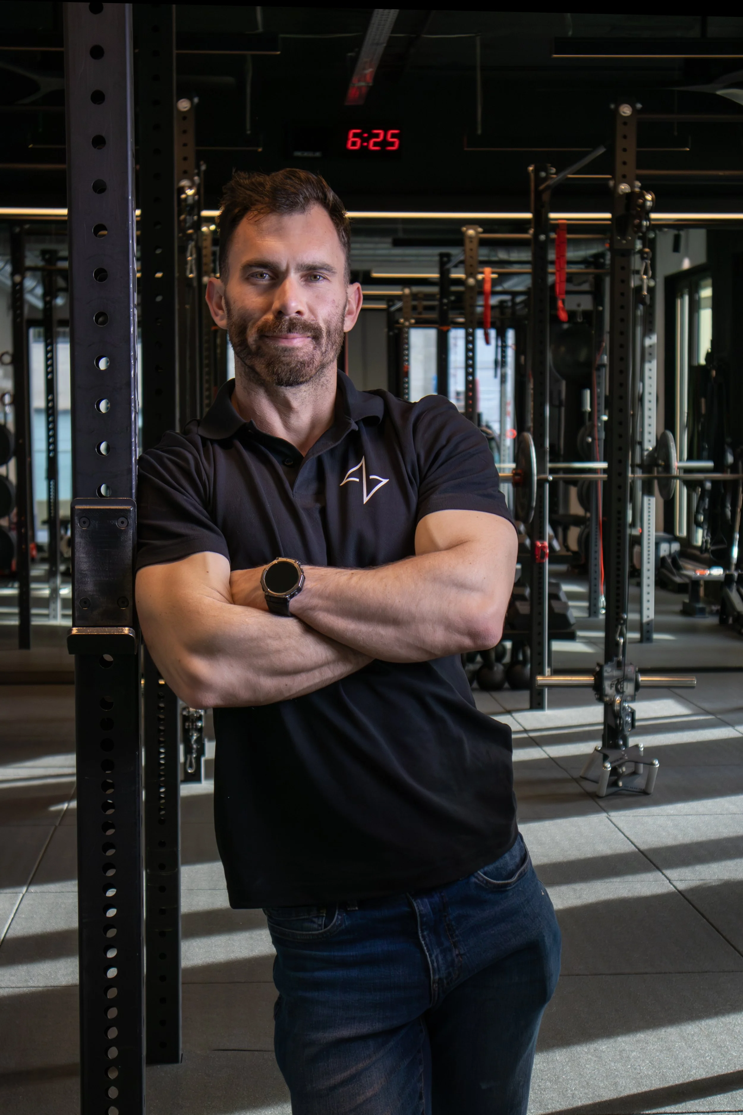 A man with dark hair and beard standing with arms crossed in a gym, wearing a black polo shirt and a smartwatch, with gym equipment and a red digital clock displaying 6:25 in the background.