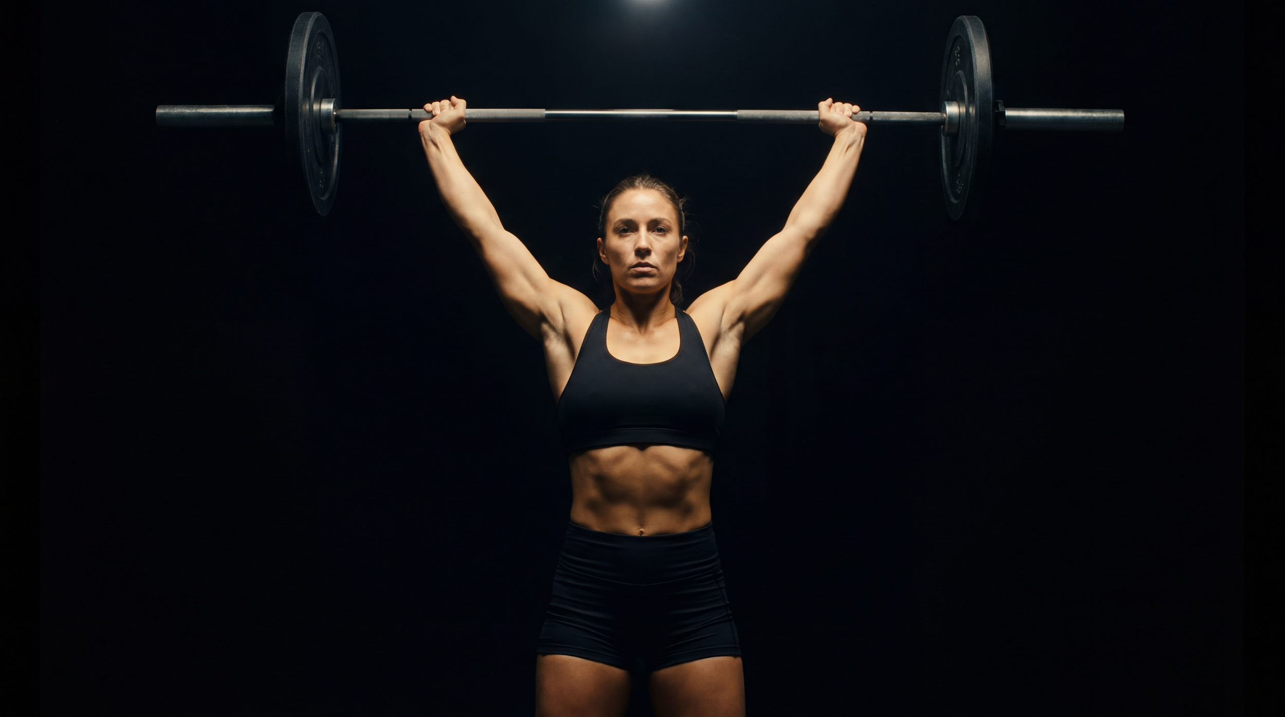 A woman lifting a barbell overhead against a black background.