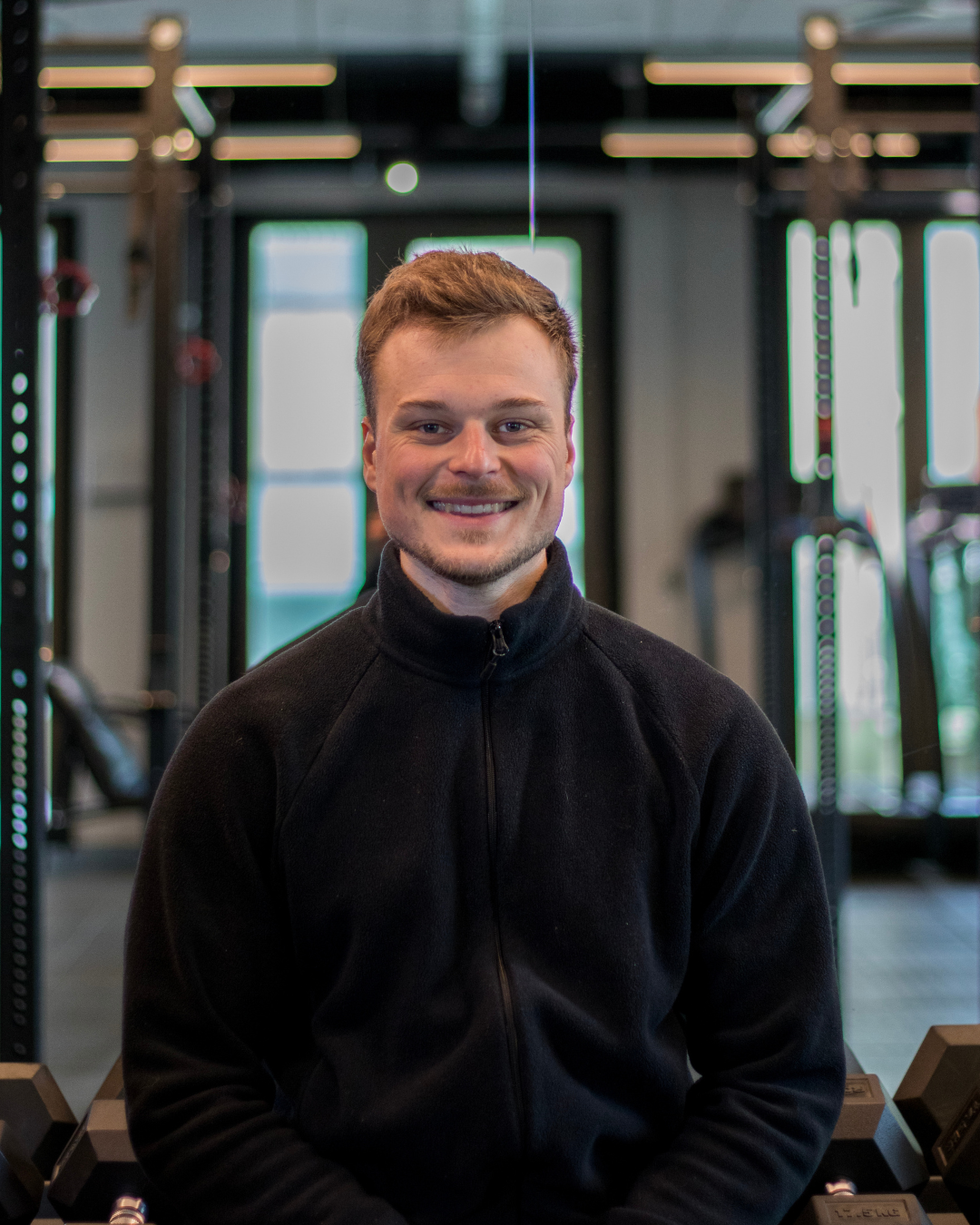 A young man with light brown hair and a beard smiling at the camera, wearing a black zip-up jacket, in a gym with workout equipment in the background.
