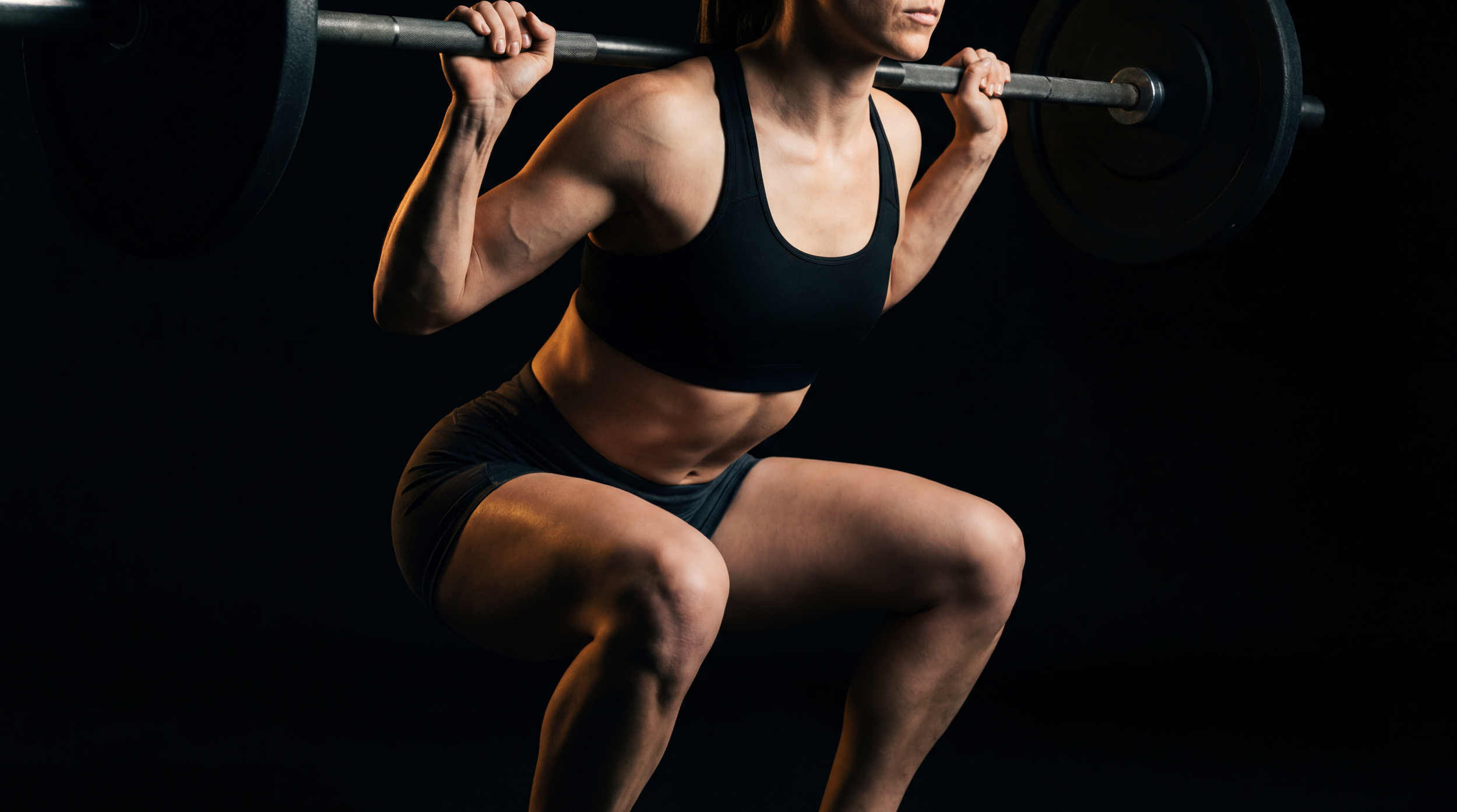 A woman in workout attire is squatting with a barbell on her shoulders in a dark gym setting.