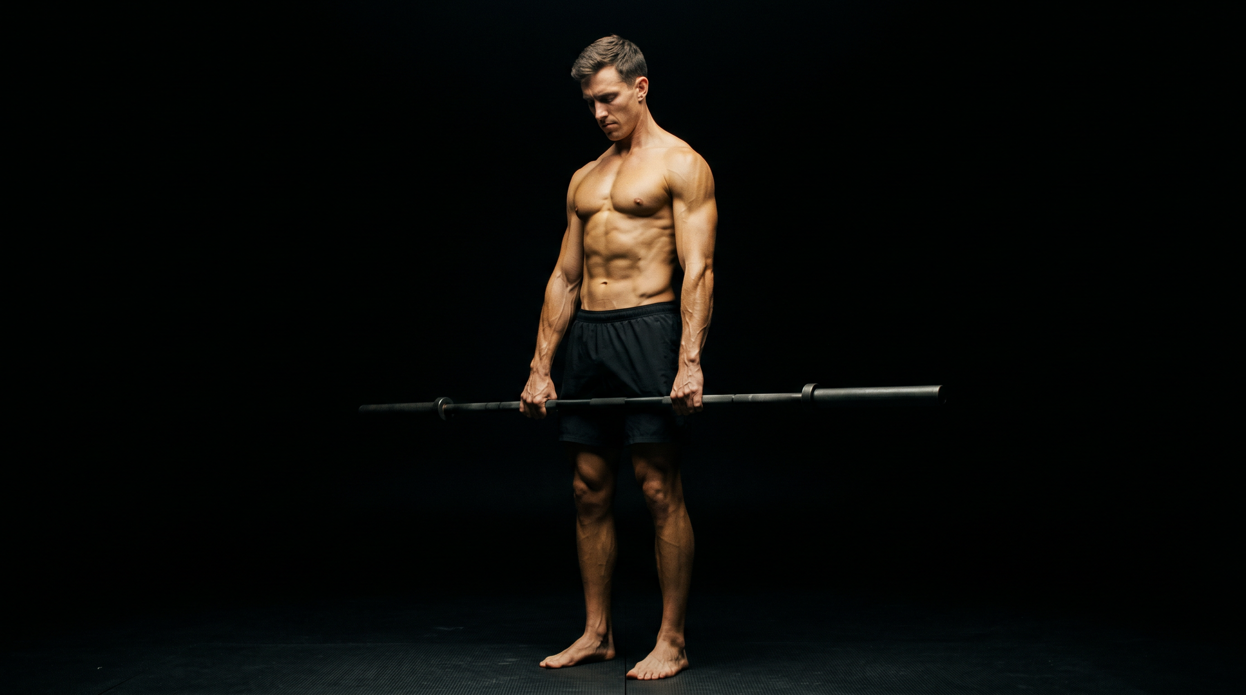 A shirtless man with a muscular build lifting a barbell in a dark studio with black background.