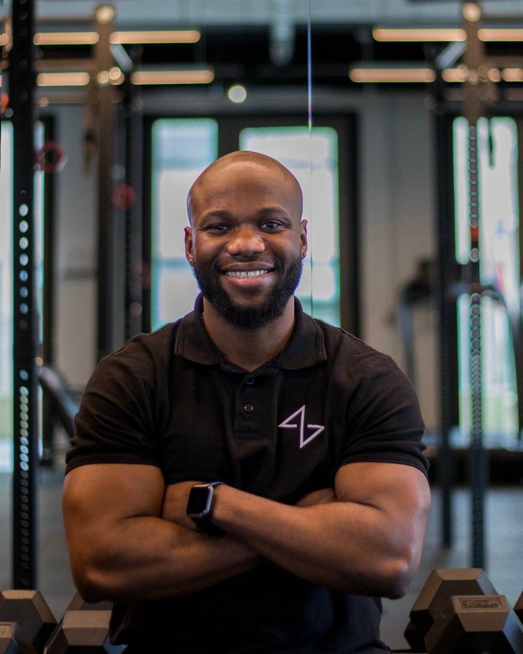 A smiling man standing with arms crossed in front of a wall with a large 'COACH' sign and a series of small photos of people in a coaching or training environment.