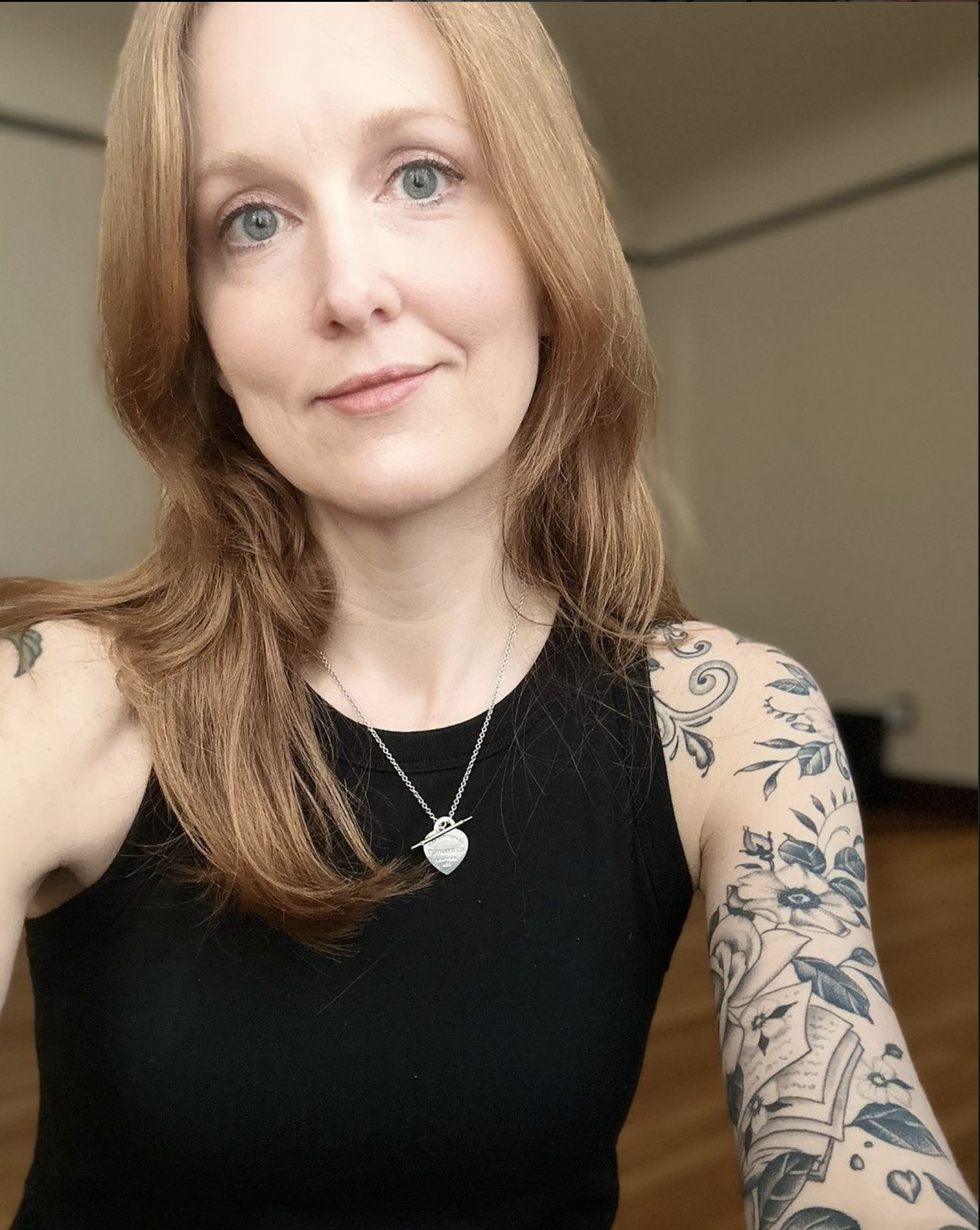 ID: A white woman with red hair and blue-grey eyes smiles at the camera while wearing a silver necklace and black tank top. There are botanical tattoos on her right arm, and she’s sitting on the floor with a wooden floor and white walls behind her.