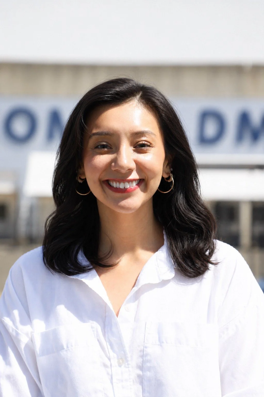 Professional headshot of a smiling woman with shoulder-length dark hair, wearing a white collared shirt, photographed outdoors.