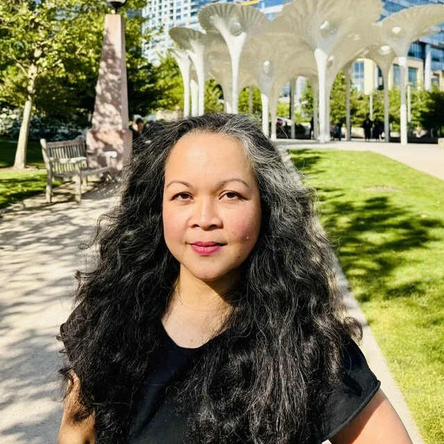 A middle‑aged Asian woman with long dark hair and a black short-sleeve shirt smiles while standing in a park.