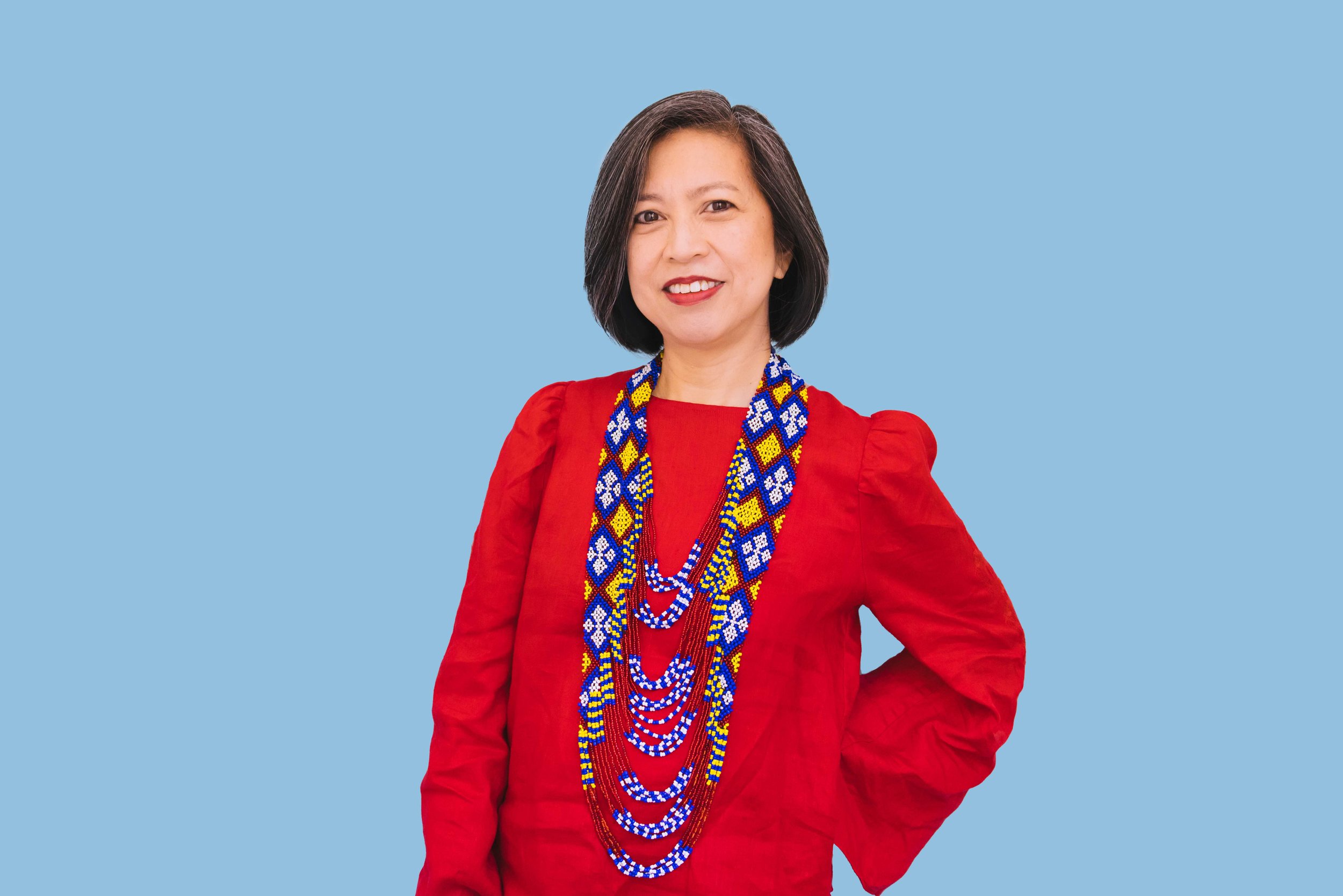 Emily P. Lawsin, a Filipina woman with light skin, straight, chin-length black hair with gray highlights, is smiling and standing with her left hand on her hip in front of a sky blue background.