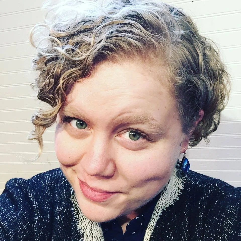 A person with short curly hair and green eyes smiles at camera