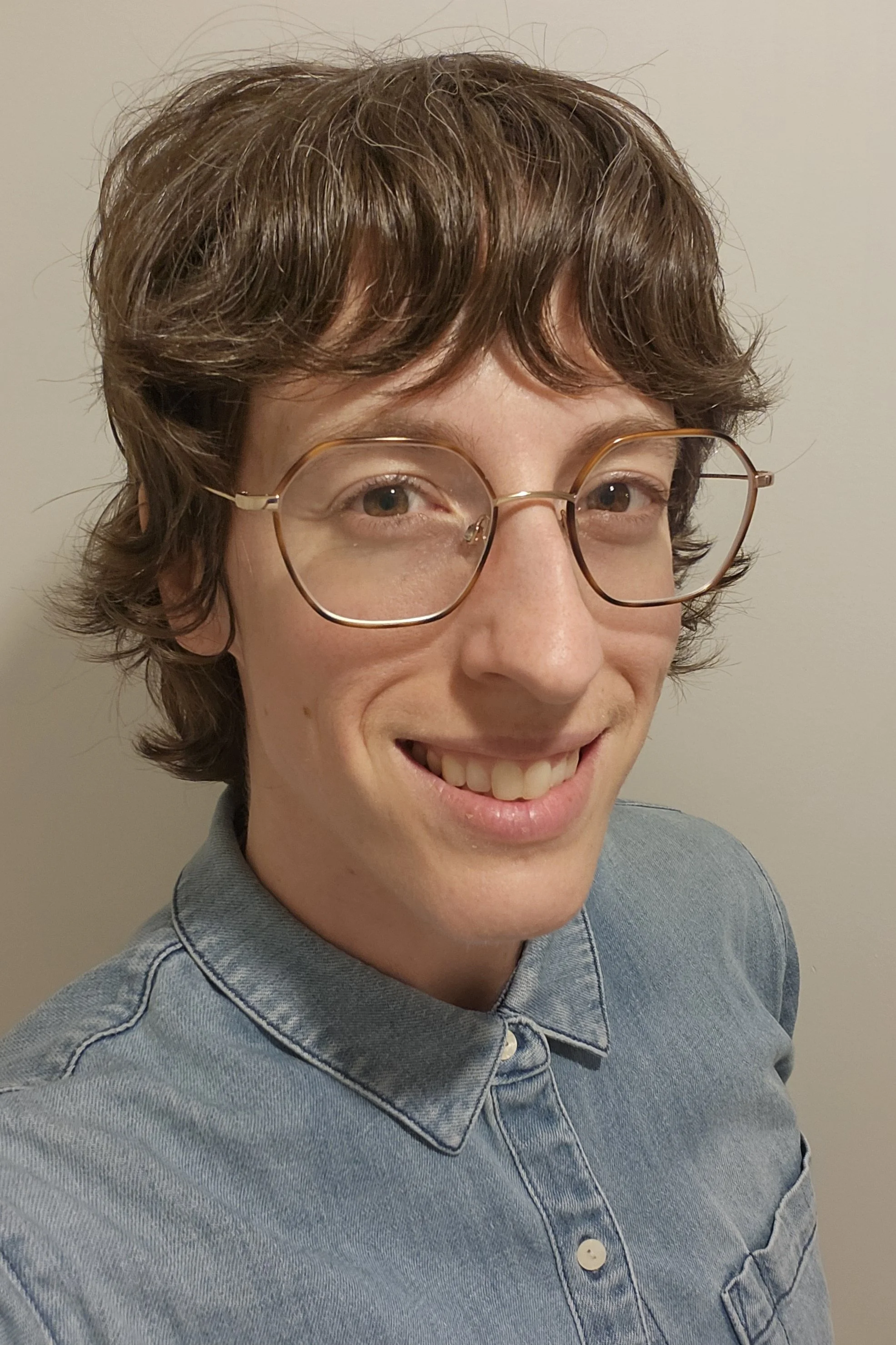 ID: A white woman with short wavy brown hair, wire-rimmed glasses, and brown eyes smiles at the camera. She wears a light denim shirt with white buttons, which are buttoned to the top of the collar.