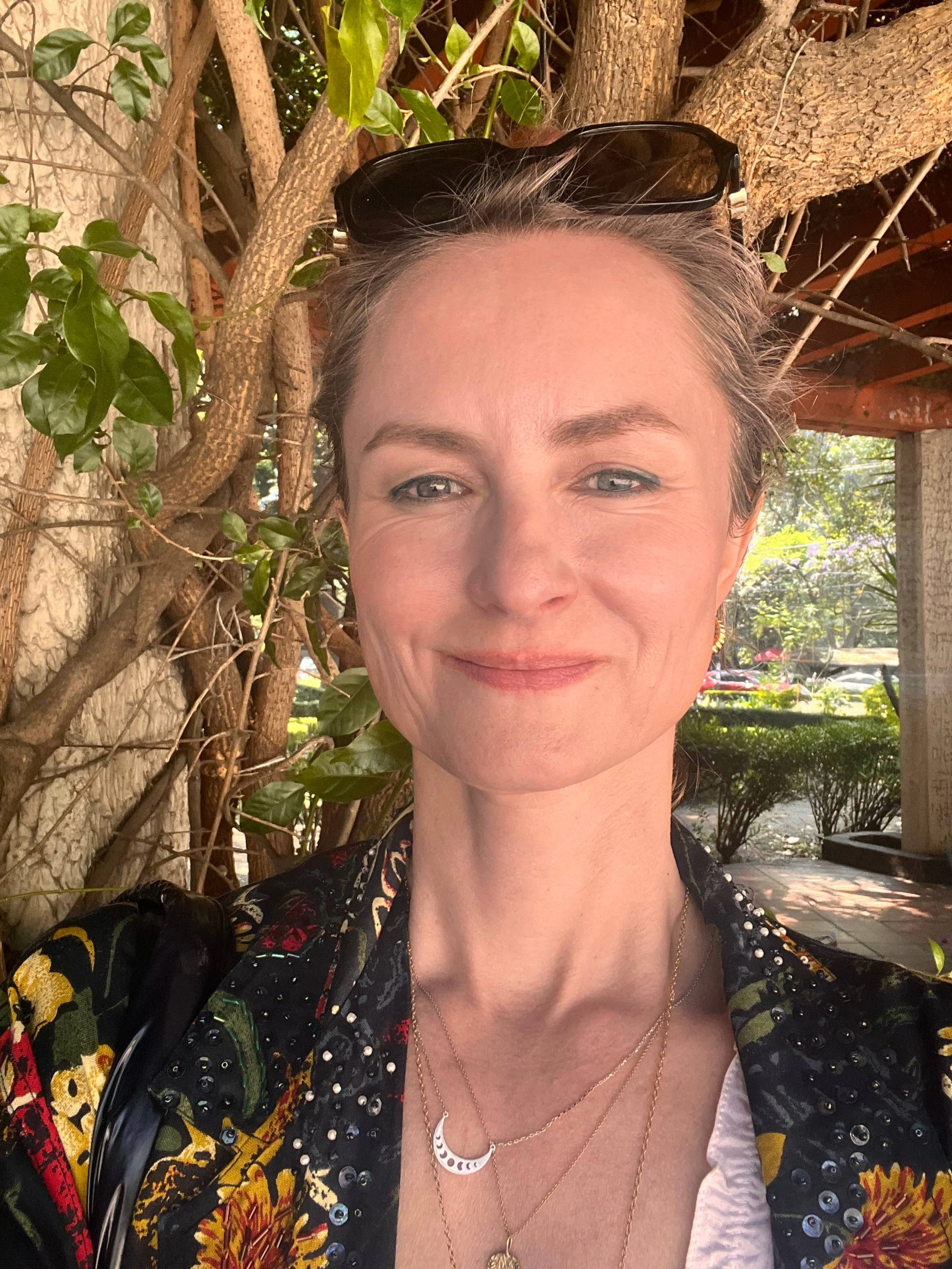 Close-up image of a woman with brown hair and grey-blue eyes smiling in front of a camera while standing in front of a tree, wearing a dark jacket, multiple necklaces, and dark brown sunglasses on her head, with a covered walkway to her right.