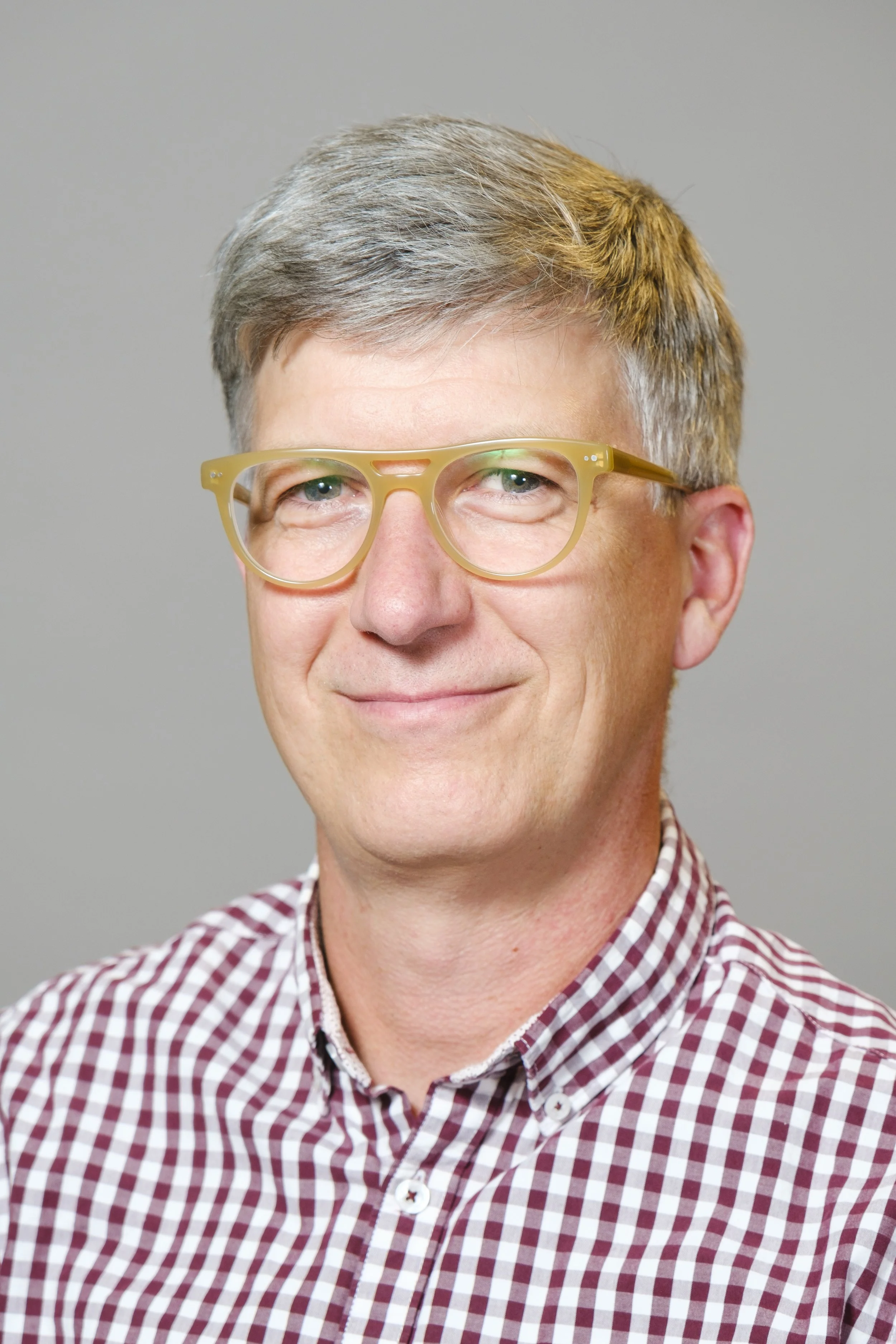 Headshot of a man with grey hair and rounded tan glasses, wearing a red-and-white checkered polo shirt in front of a light grey background.