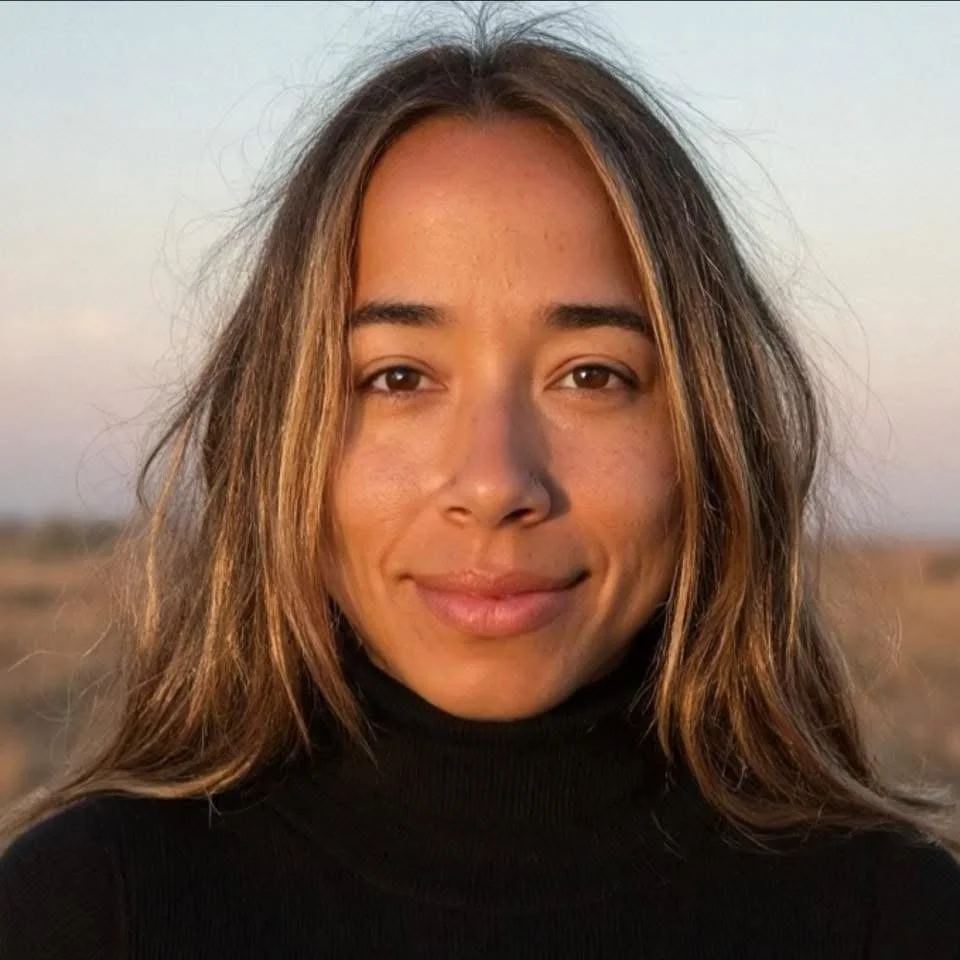Close-up portrait of a smiling woman with long brown hair in an outdoor setting during sunset.