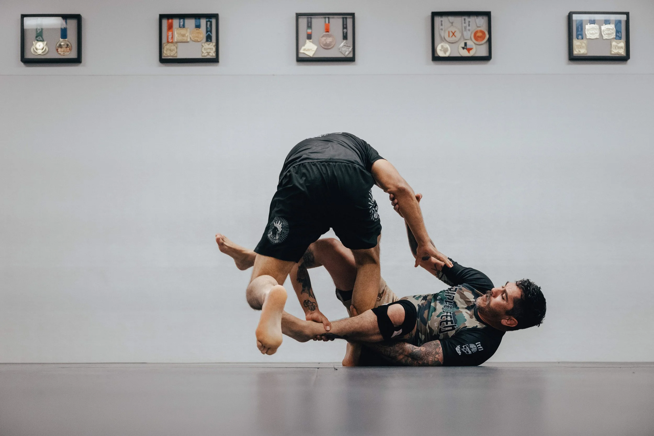 Two men practicing No-Gi Brazilian Jiu-Jitsu, one in a black rash guard executing a move on the other, who is in camouflage shorts and lying on the floor, in front of a white wall with framed medals displayed at Carpio Jiu Jitsu in Long Beach