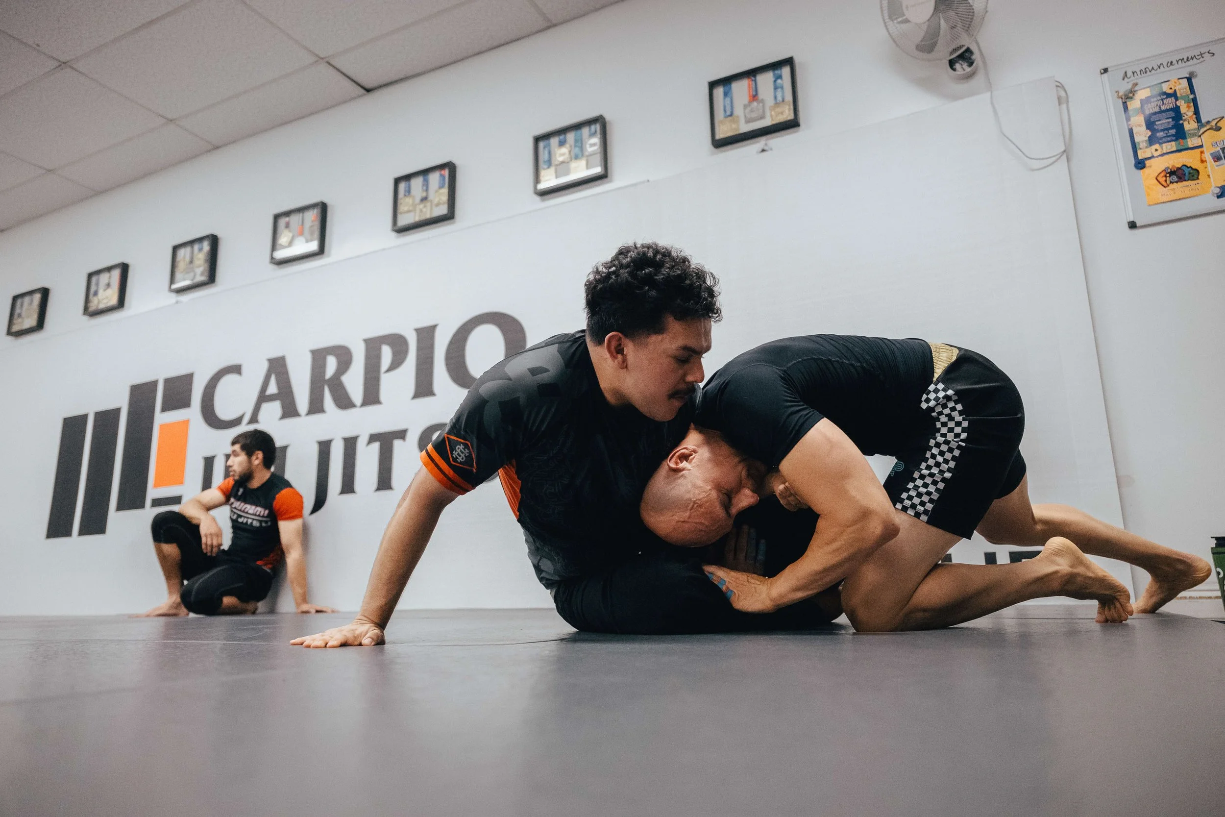 Martial arts class practicing No-Gi grappling techniques with two men engaged on the mat, while another person observes in the background at Carpio Jiu Jitsu in Long Beach.