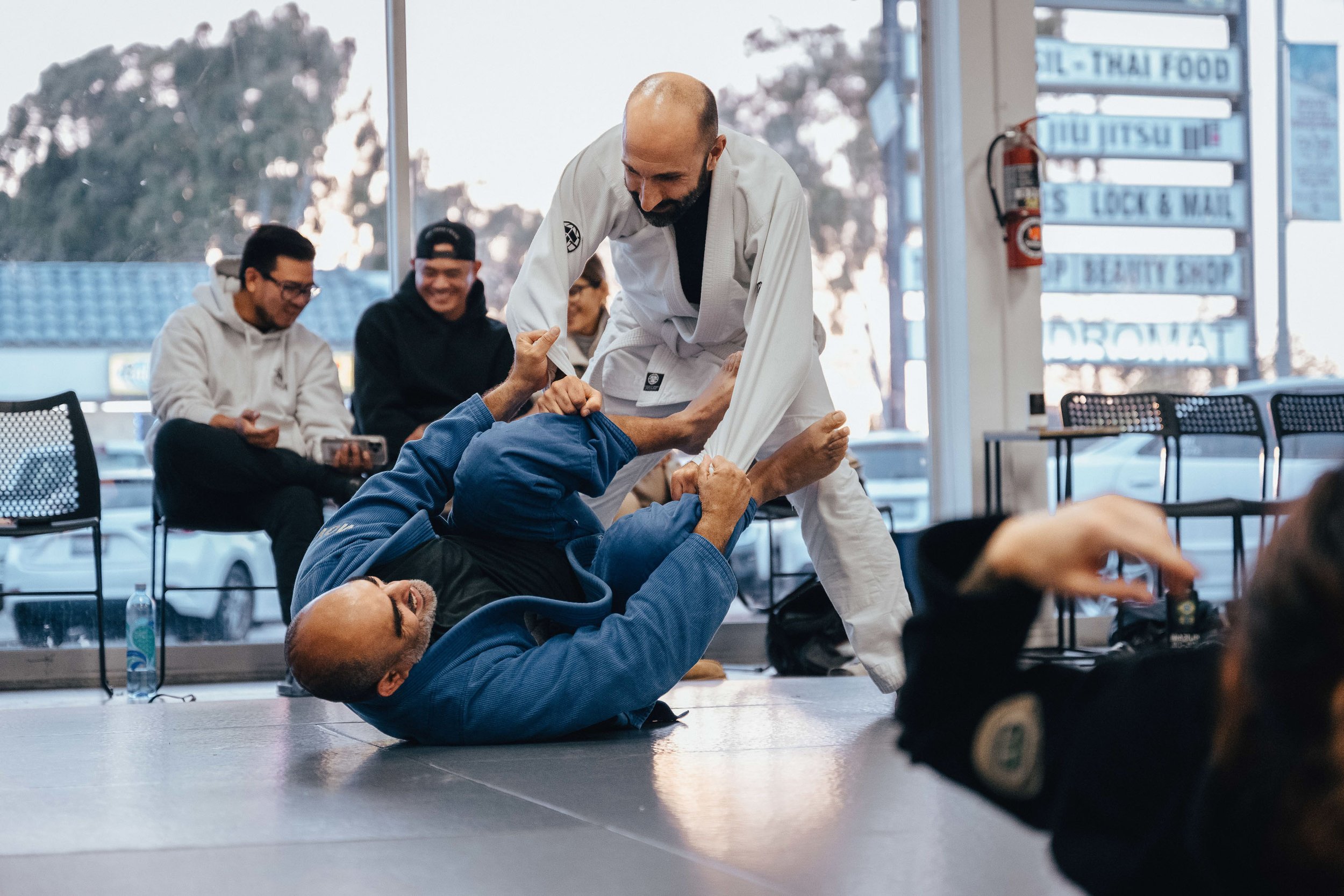 People practicing Brazilian Jiu-Jitsu in a martial arts training room with large windows, some sitting and smiling, while one person in a blue gi is on the ground gripping another person's arm.