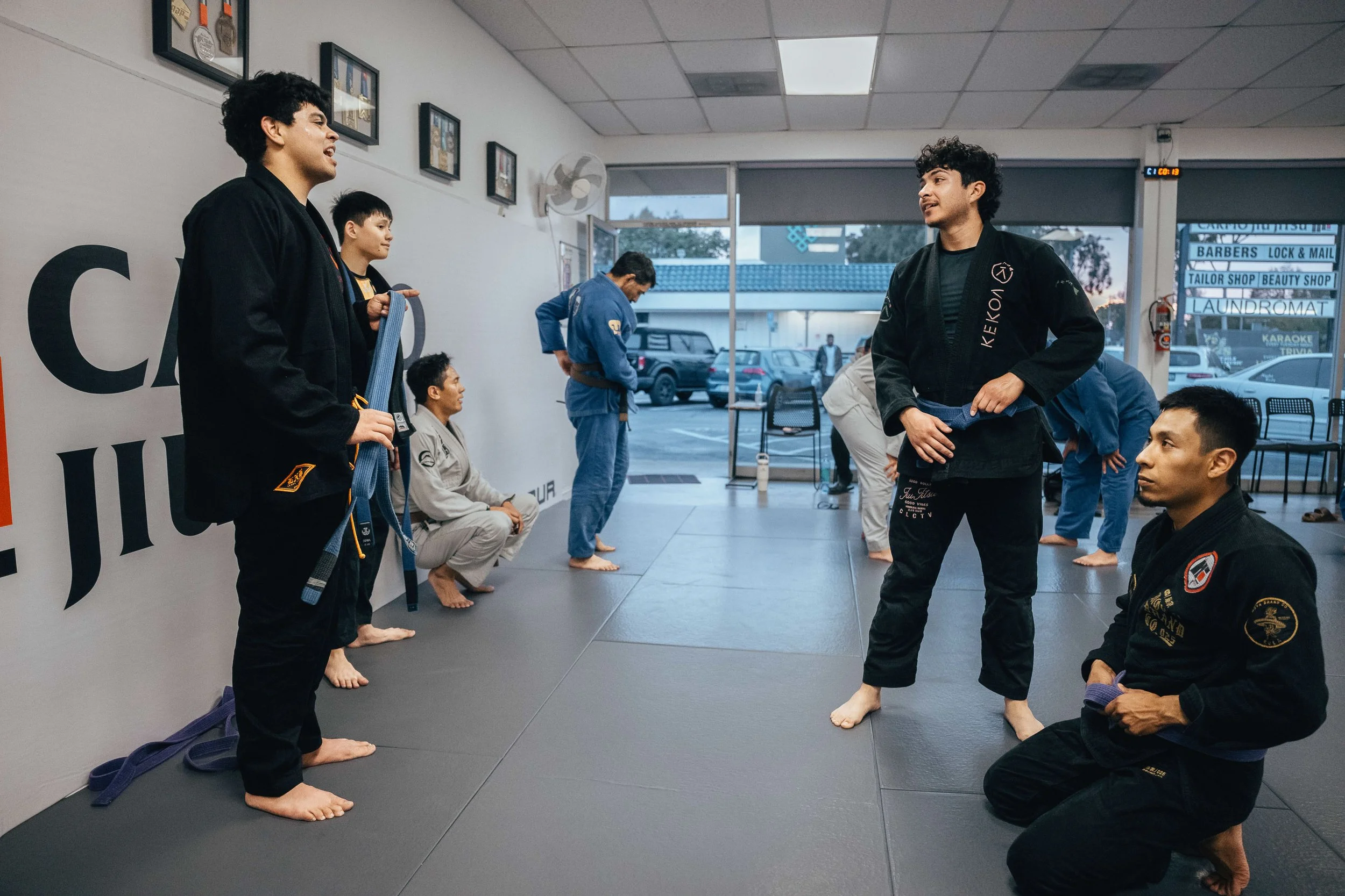 Martial arts students in black uniforms listening to instructor in a dojo with posters on the wall and cars outside through large windows.
