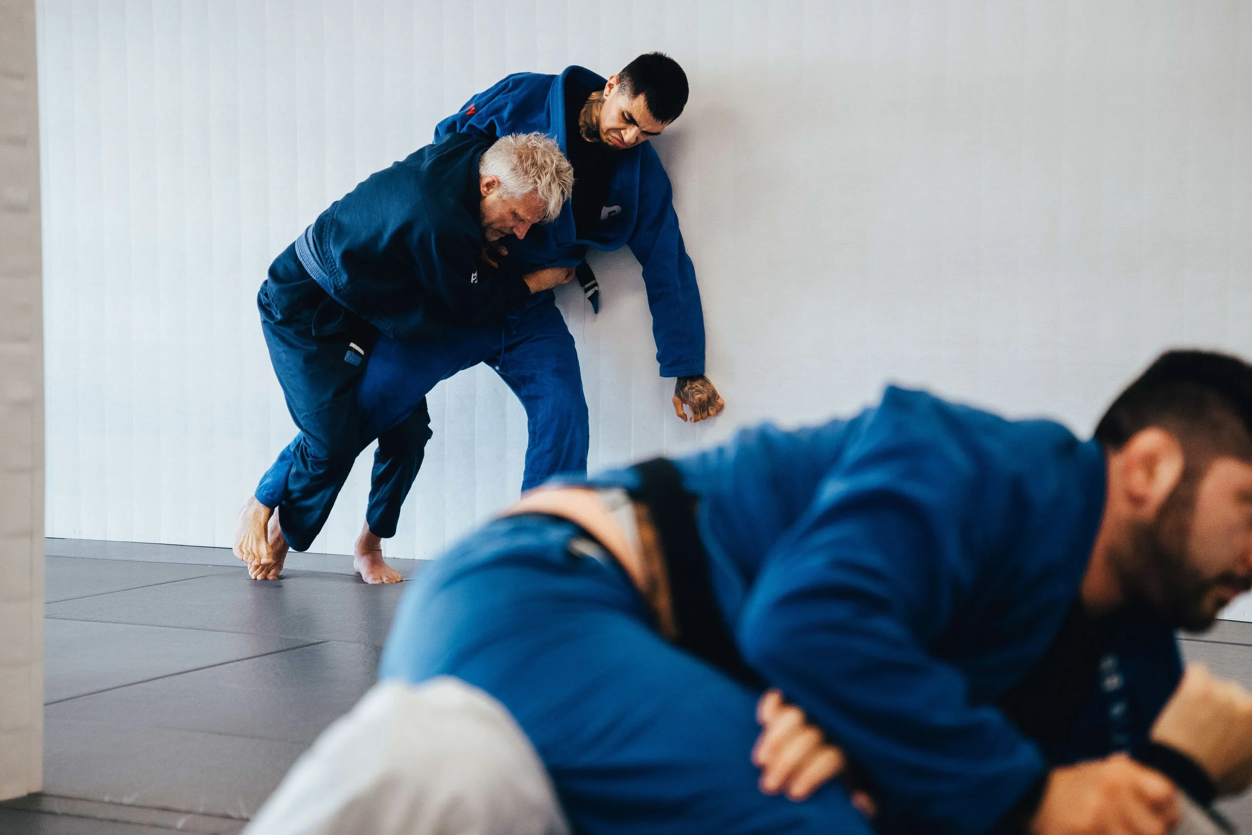 Two men wearing blue gis practicing Brazilian Jiu-Jitsu on a gray mat at a martial arts gym. One man with blond hair is passing the guard of another man with dark hair, both focused and engaged in the training session.