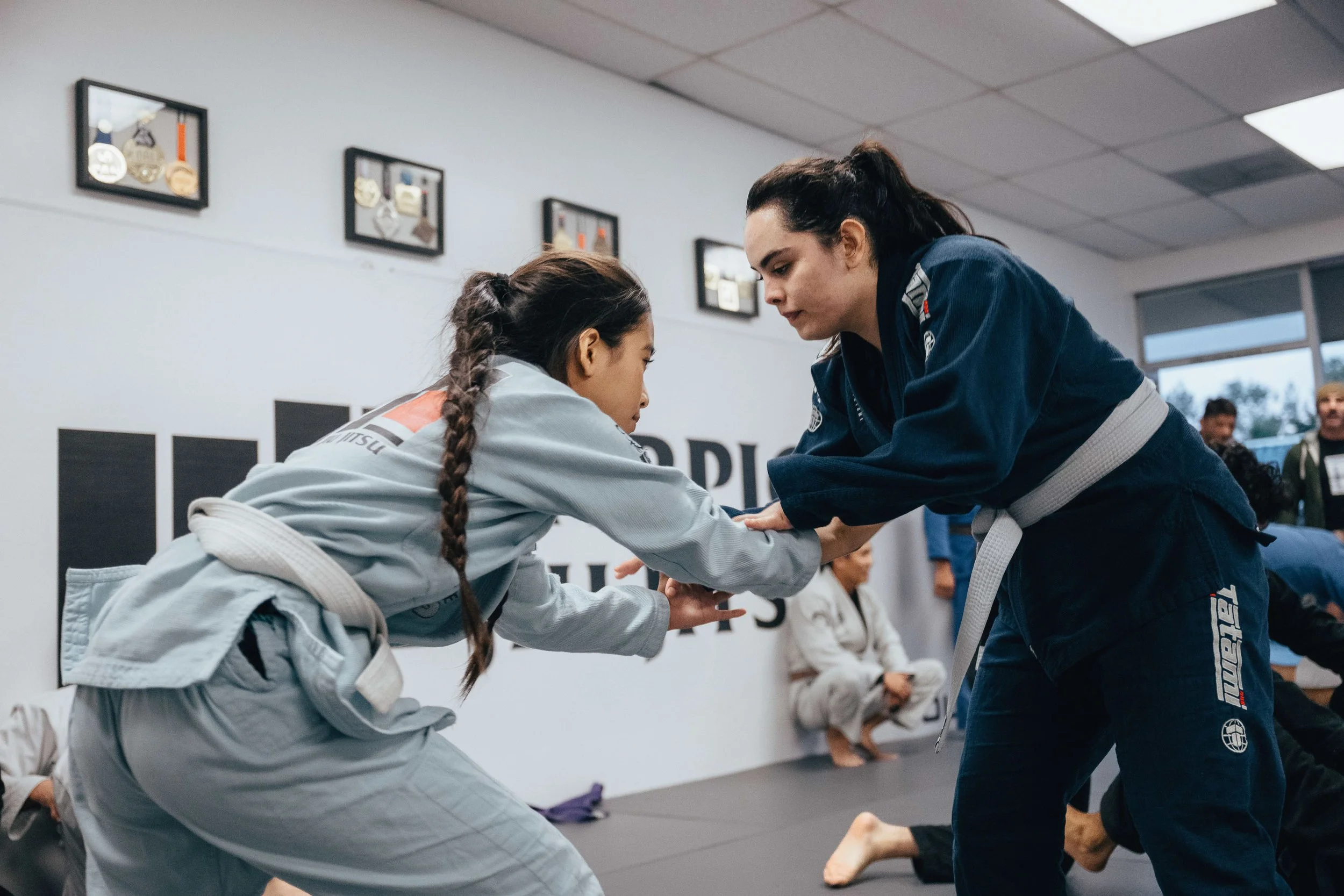 Two women practicing Brazilian Jiu-Jitsu on a training mat. One woman is in a white gi and the other in a dark blue gi, engaging in a grappling move.