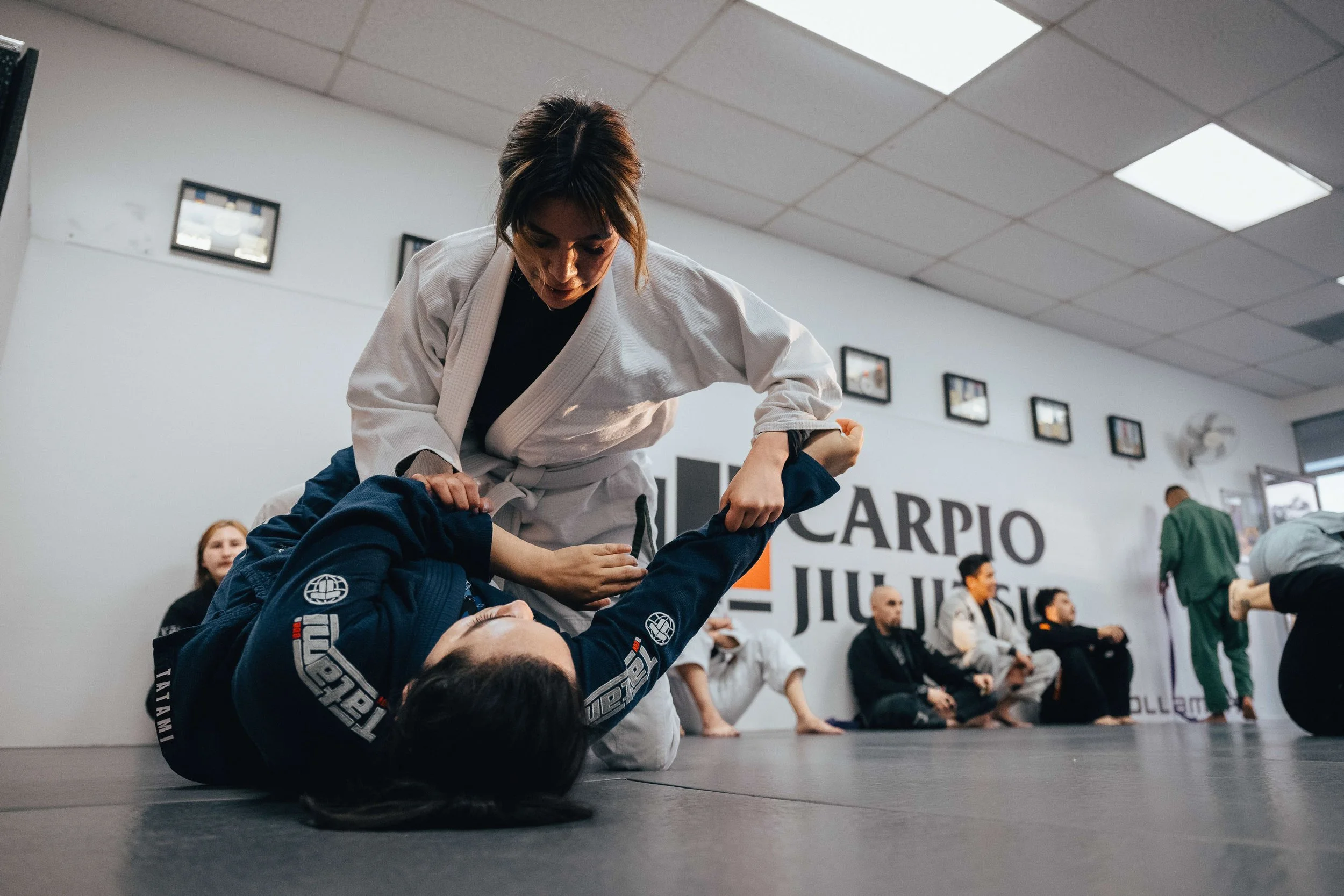 Martial arts class with a woman in a white gi demonstrating a technique on a man in a black gi, in a gym with other students and instructors observing.