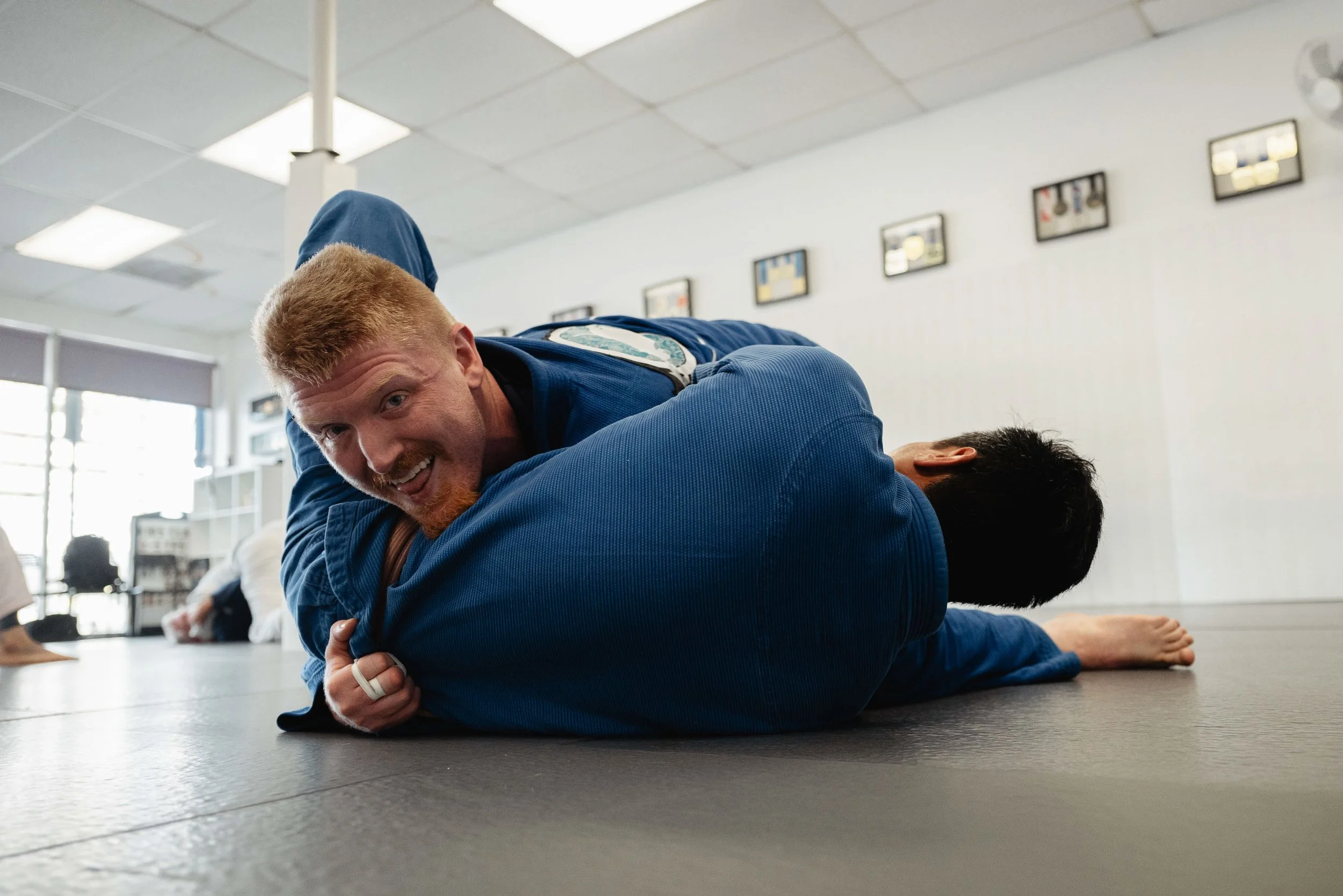 Two martial artists practicing Brazilian Jiu-Jitsu on a mat, with one person in a blue gi applying a technique on another person in a blue gi, in a well-lit indoor training facility.