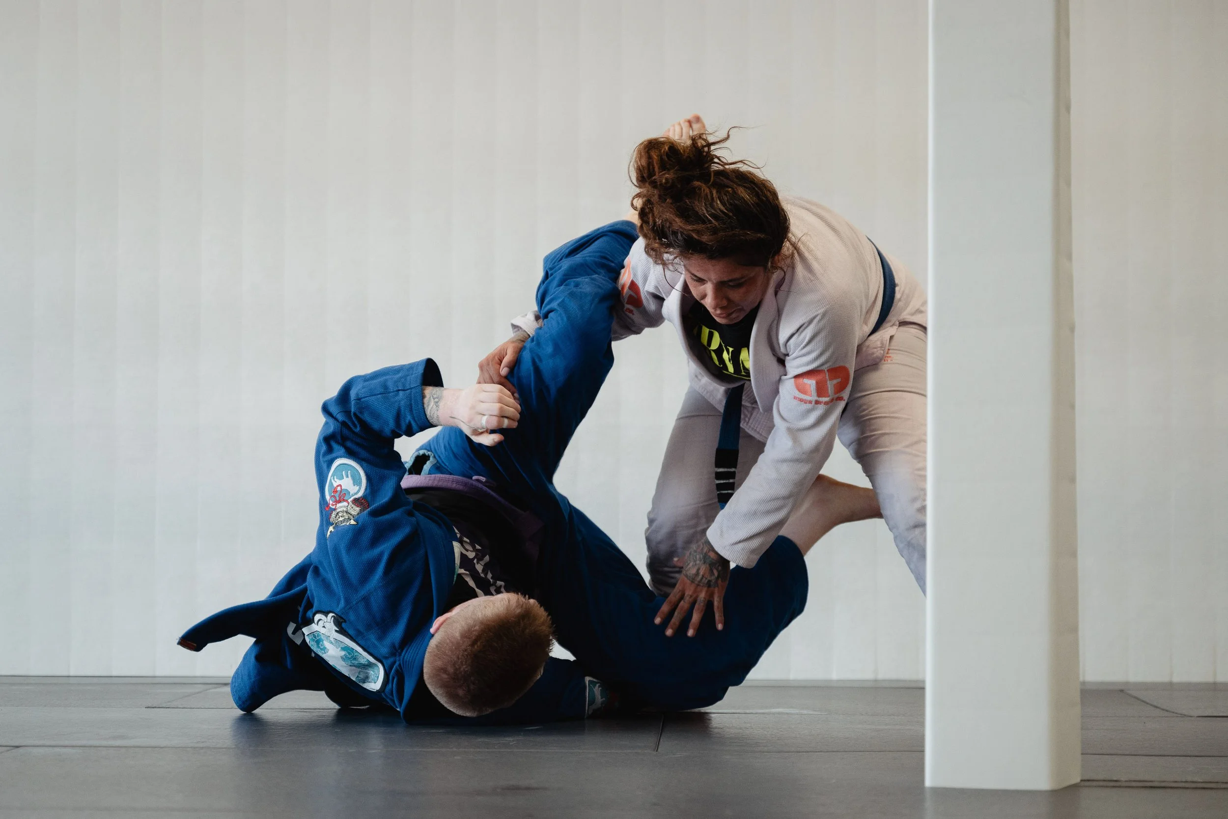 Two women practicing Brazilian Jiu-Jitsu on the floor of a training facility. One woman in a blue gi holds the other woman, who is in a white gi, in a grappling position. The woman in white is attempting a submission move while the woman in blue is d