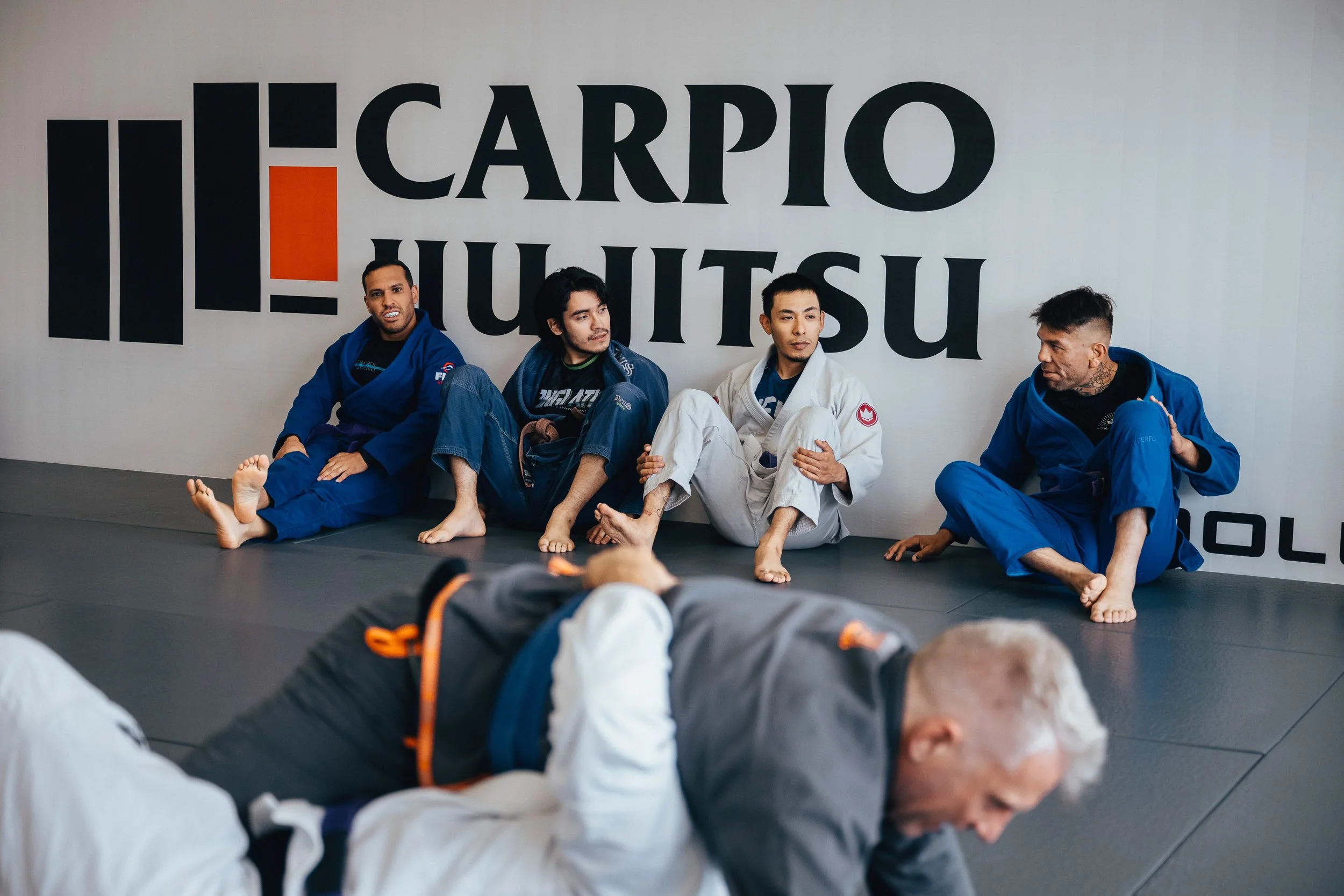 Four martial artists sitting against a wall with the words "CARRJO JIU JITSU" on it, dressed in gi uniforms, inside a martial arts gym or dojo in Long Beach