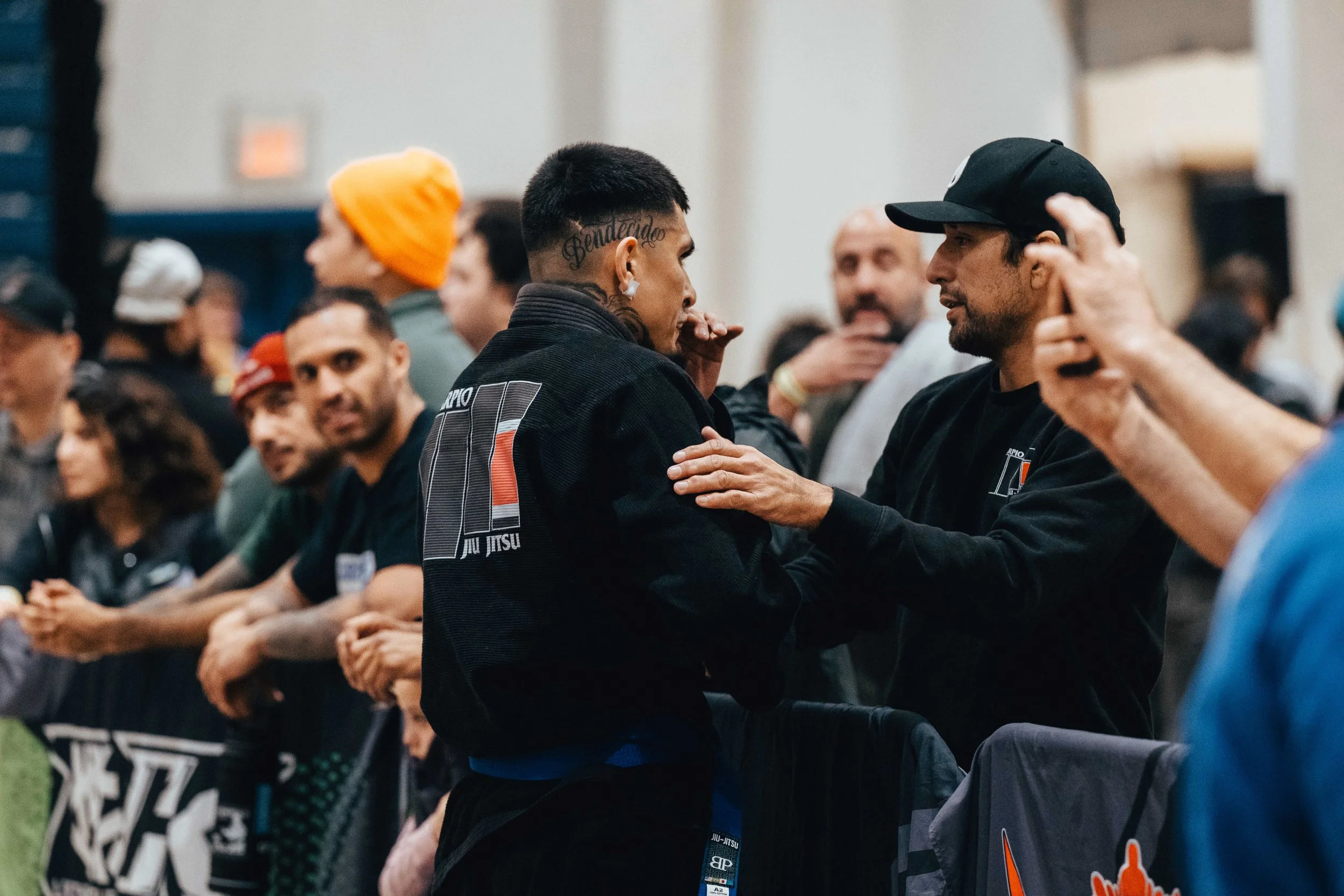 Two men with similar black T-shirts engaging in a conversation at a crowded indoor event, likely martial arts or sports exhibition.