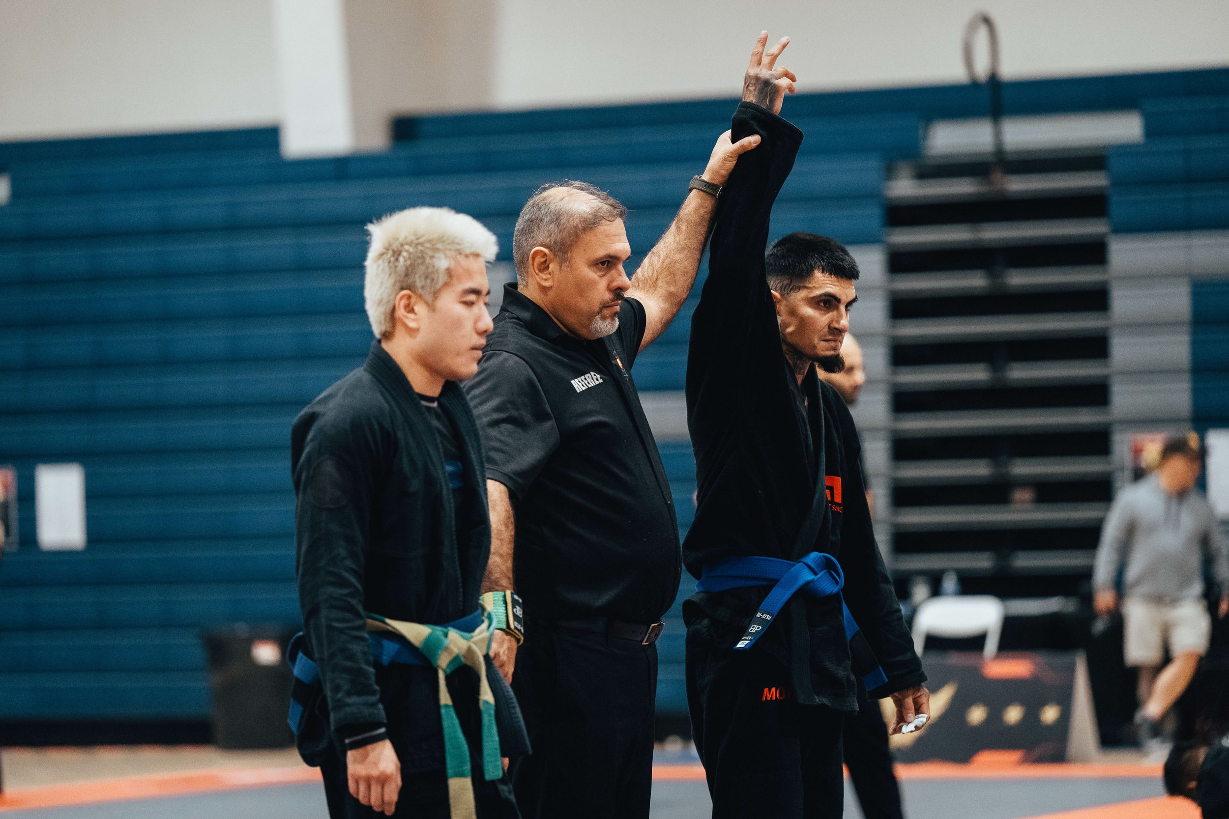 Three martial artists standing in a line at an indoor gym, with one man raising his hand. All are wearing black gi uniforms with blue belts, and the man raising his hand has a tattoo on his neck and a small tattoo on his forearm.