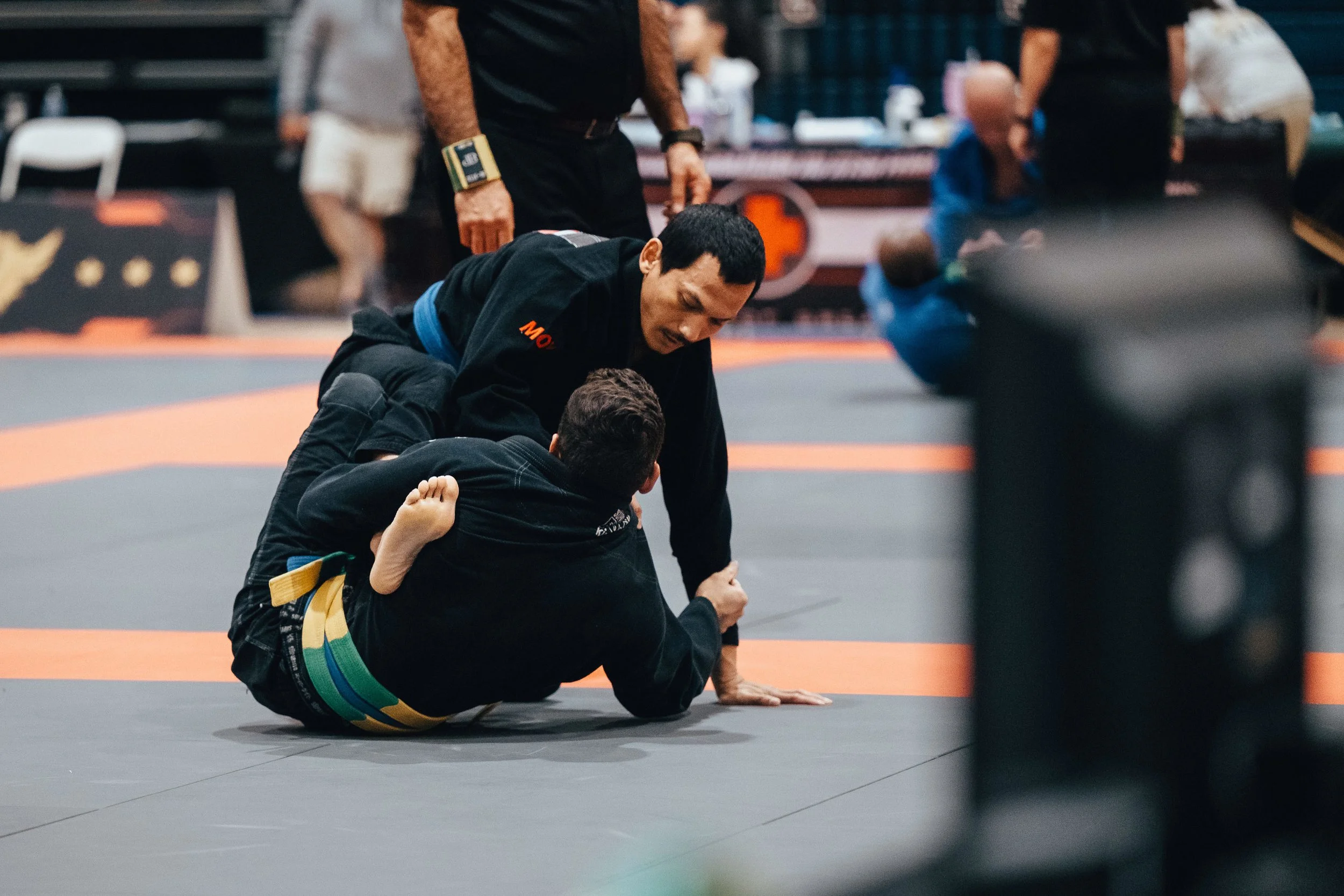 A man with short black hair and wearing a black jiu-jitsu gi is on the floor, holding a young boy with short dark hair and a black gi with colorful belt, in a grappling position during a Brazilian Jiu-Jitsu training or competition. Several people and