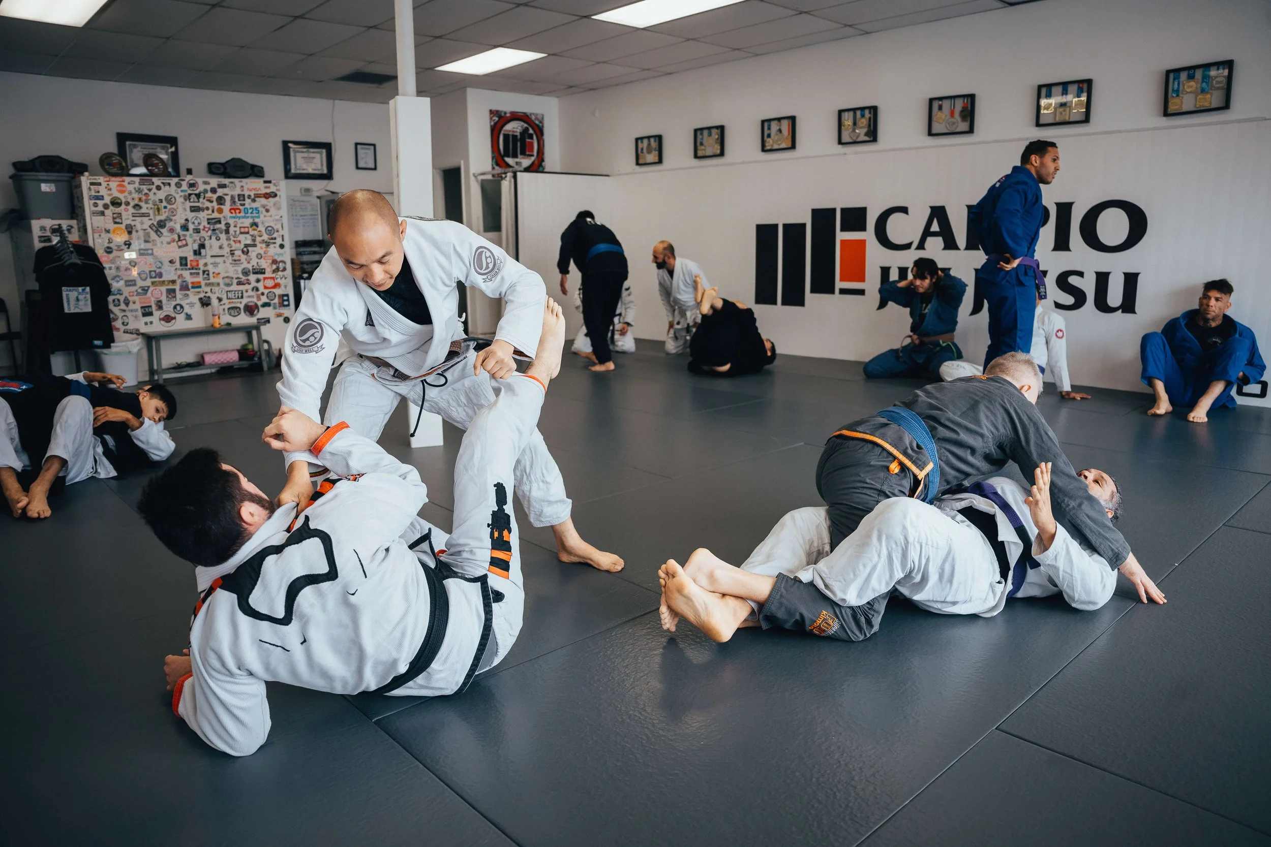 Martial arts class practicing techniques on a black mat in a gym with several students wearing gi uniforms and a large logo on the wall that reads 'CaeiO Jiu Jitsu'.