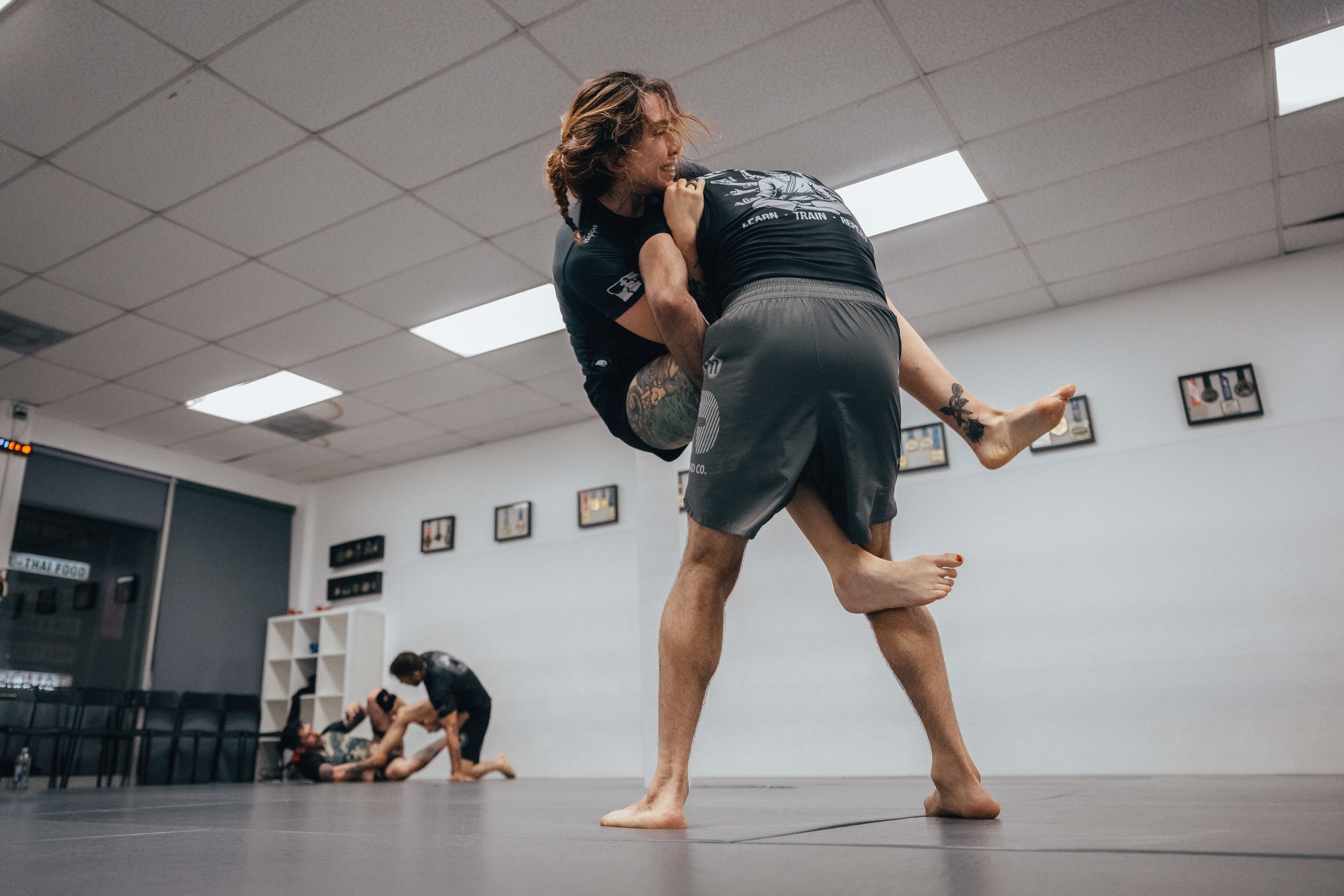 Two individuals practicing No-Gi Brazilian Jiu-Jitsu on a mat in a martial arts gym, with one person holding the other in a grappling position at Carpio Jiu Jitsu in Long Beach