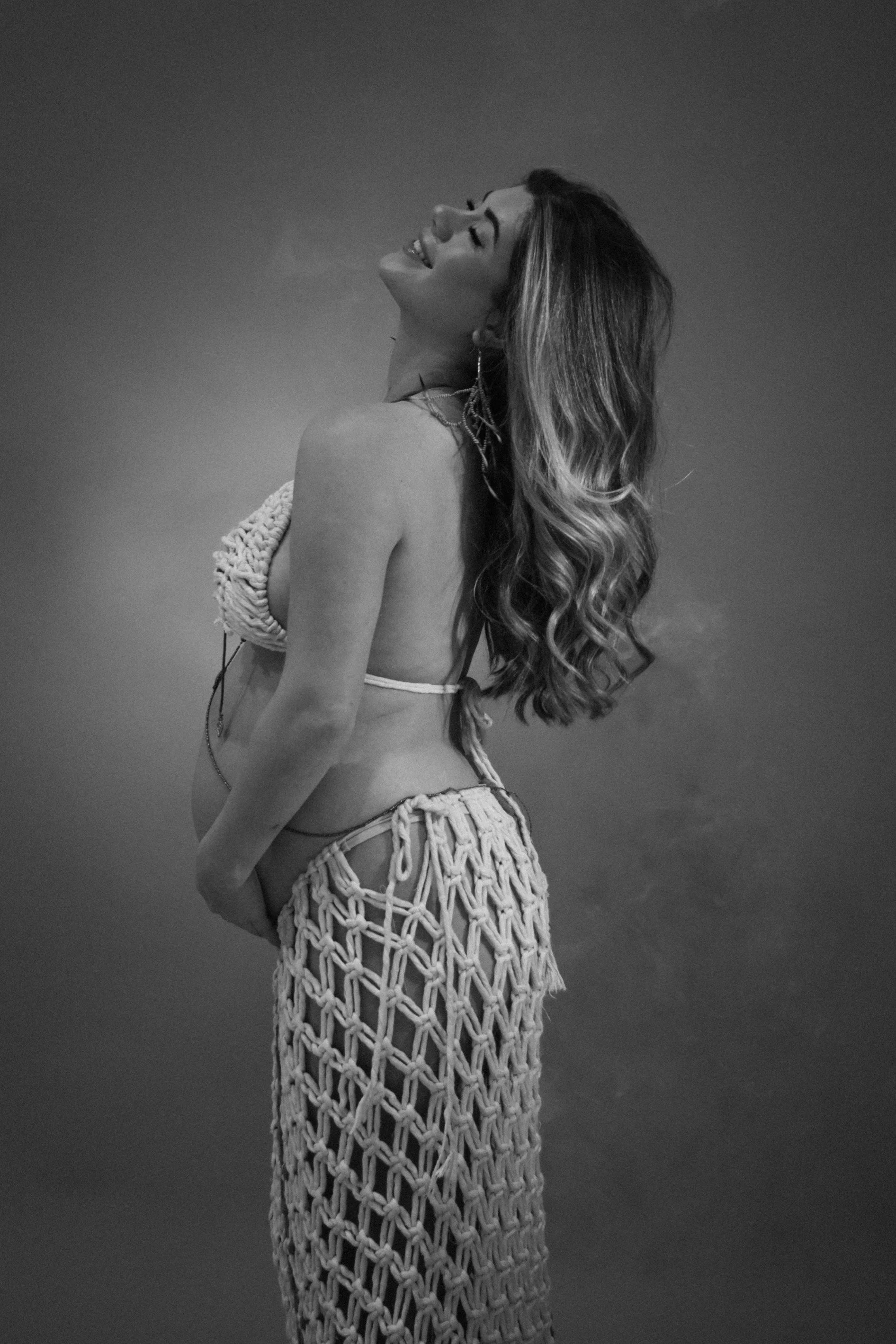 A woman with long wavy hair smiling with her head tilted back, standing outdoors with a cloudy sky in the background.